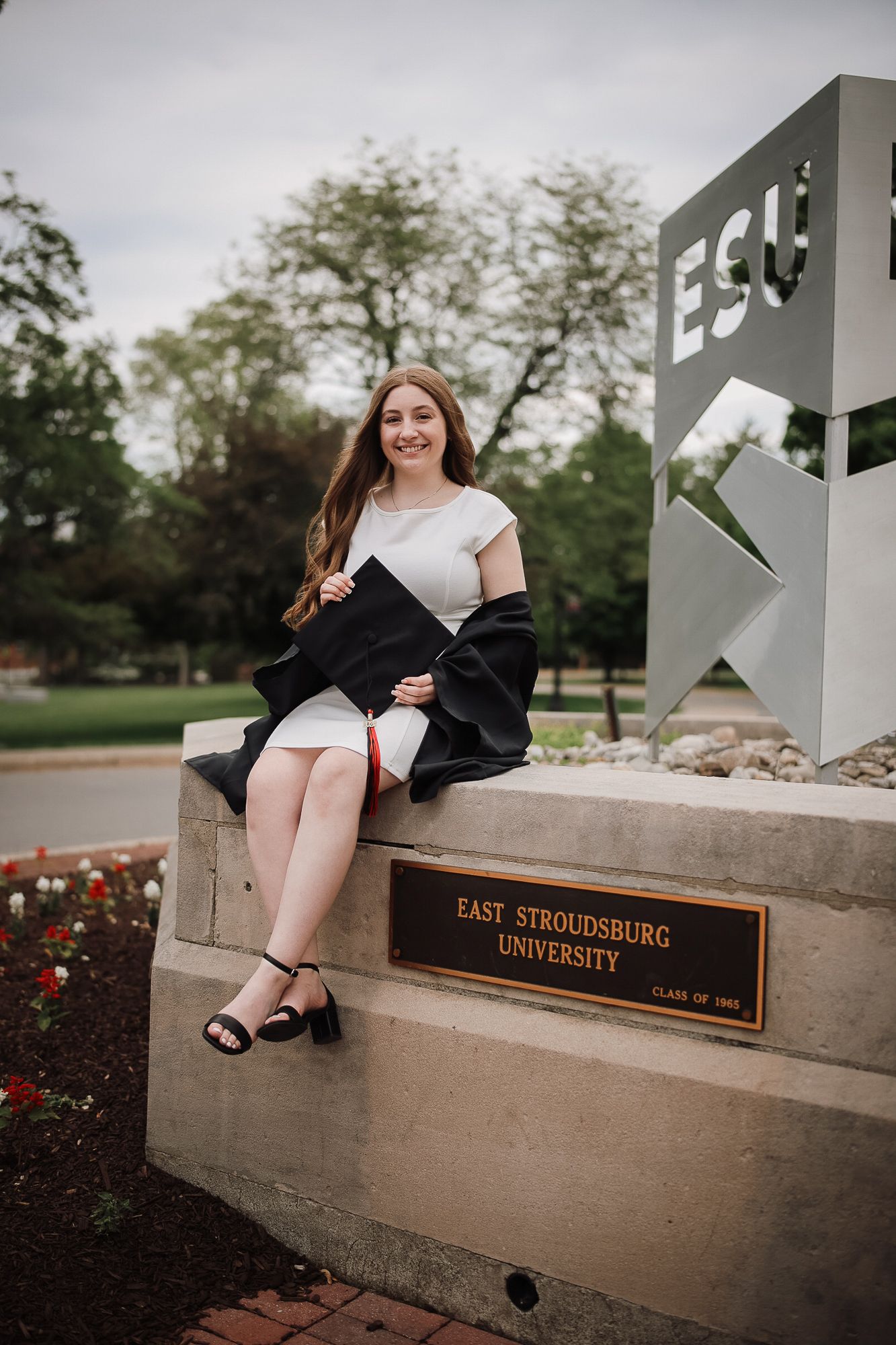 A smiling graduate in a white dress holds her cap beside the East Stroudsburg University sign on campus.