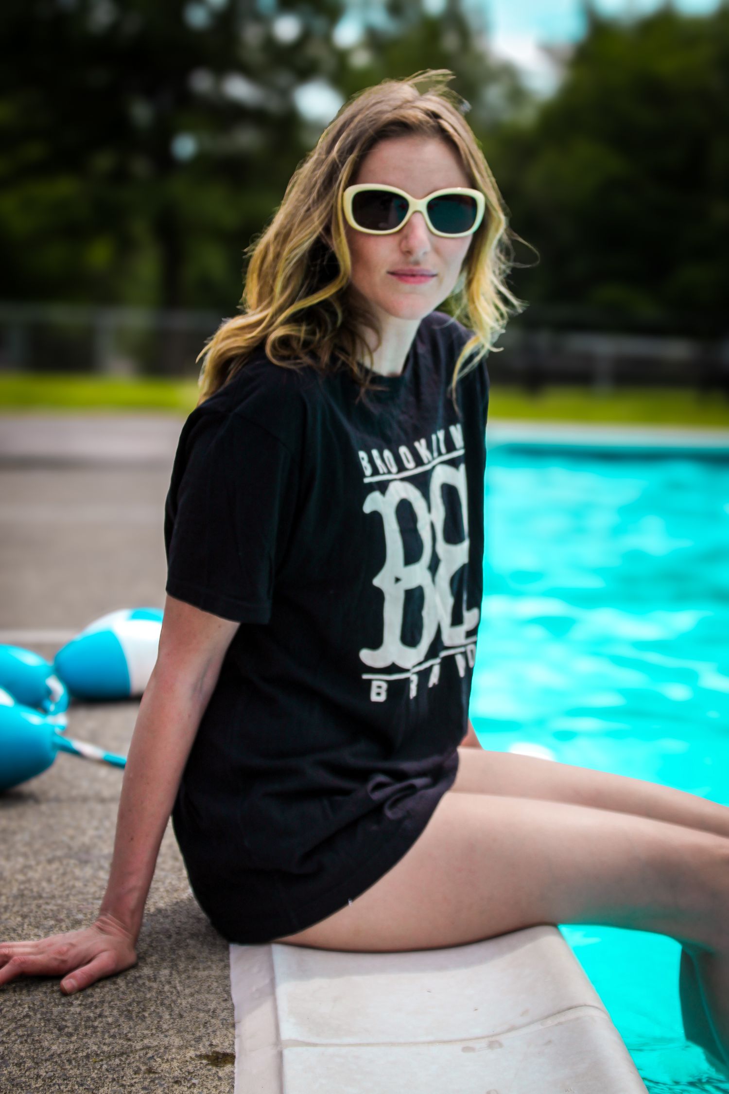 Person in black t-shirt lounging by bright turquoise pool wearing sunglasses on sunny summer day.