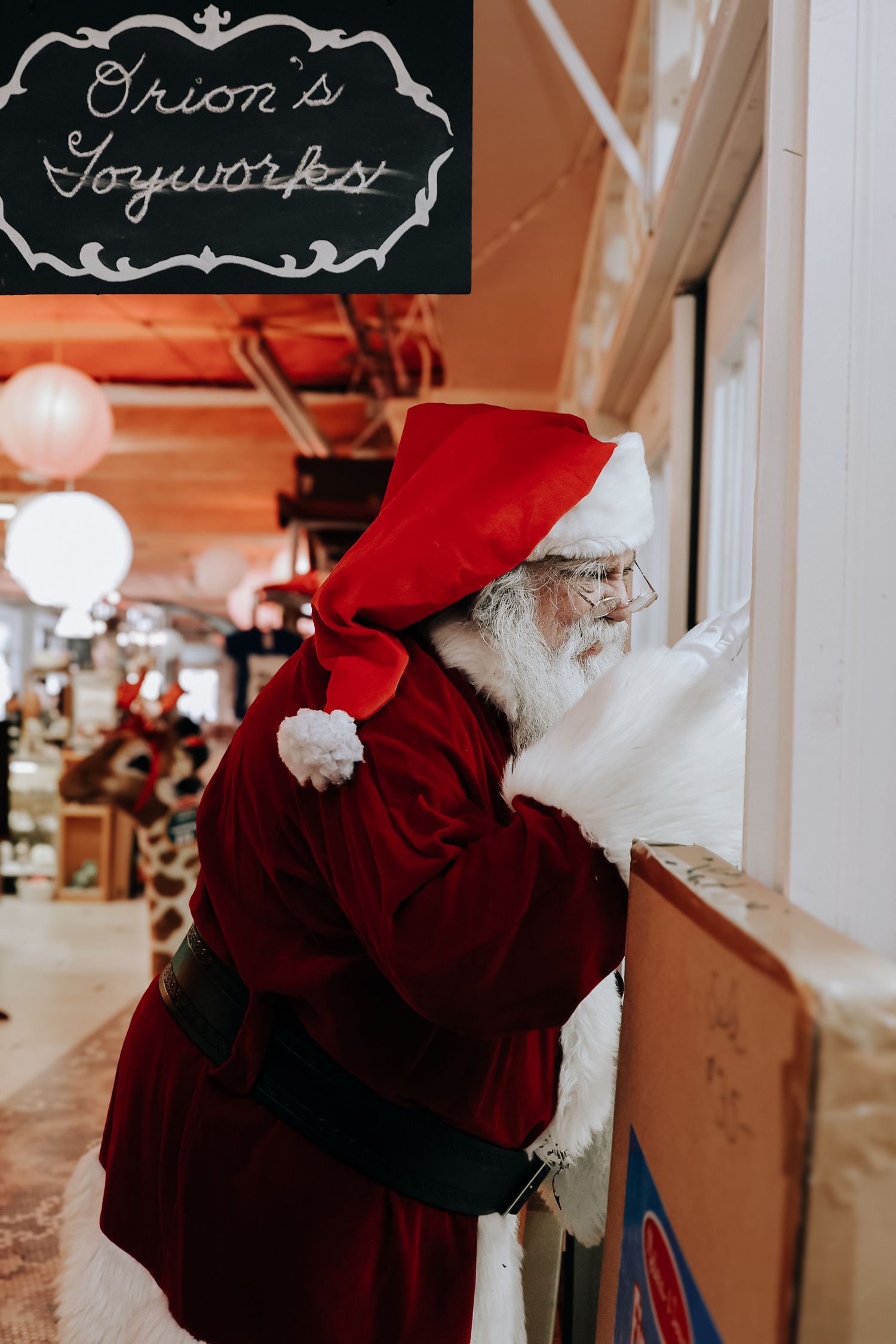 A festive figure in red and white stands in a holiday-themed indoor setting with chalkboard signs visible in the background.