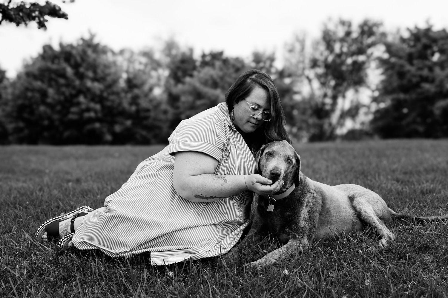 A tender moment between a person in a dress and their beloved dog laying together on grassy field in black and white.