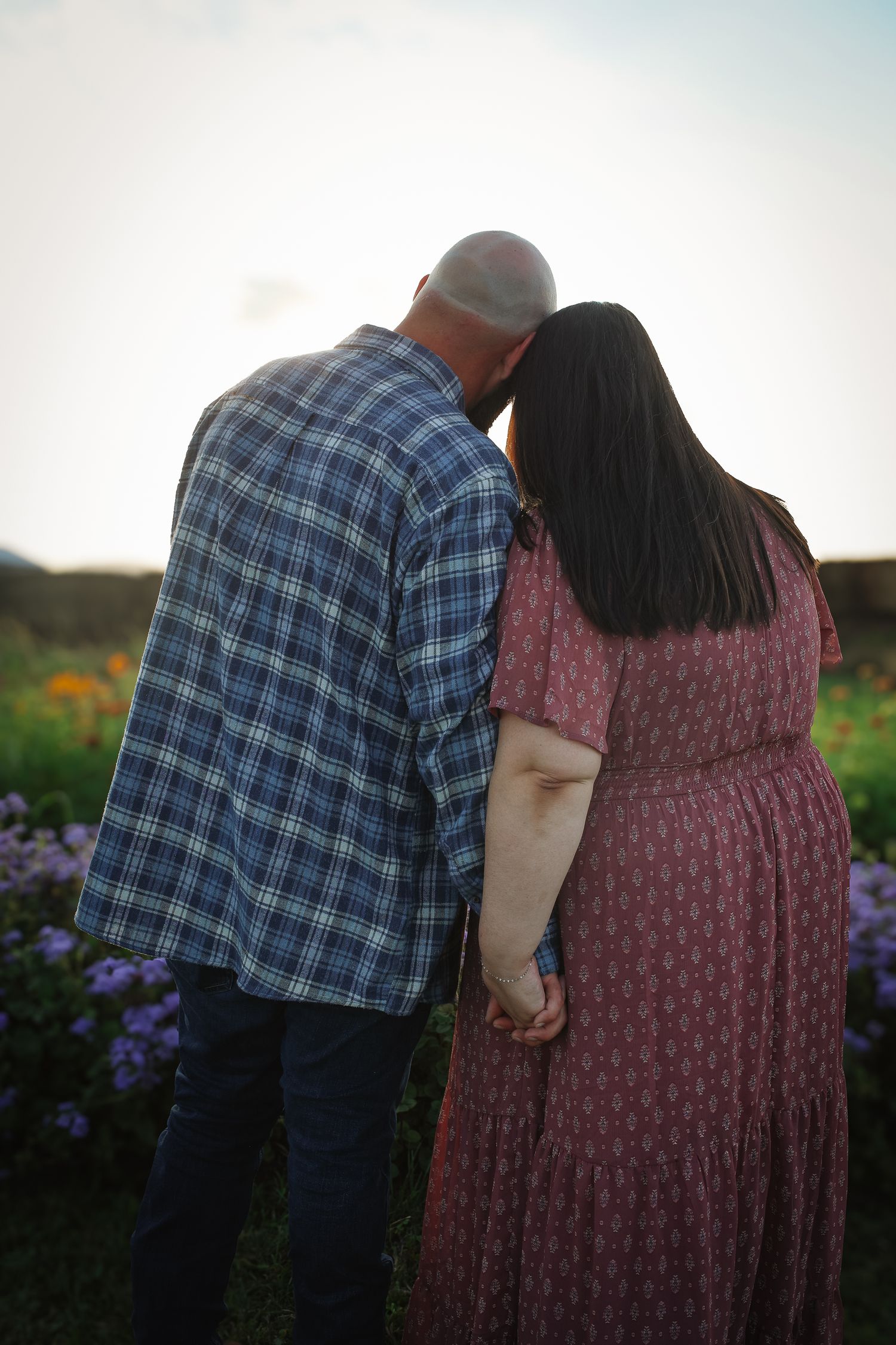 A couple in casual attire stands together in a garden at sunset, sharing an intimate moment among blooming flowers.