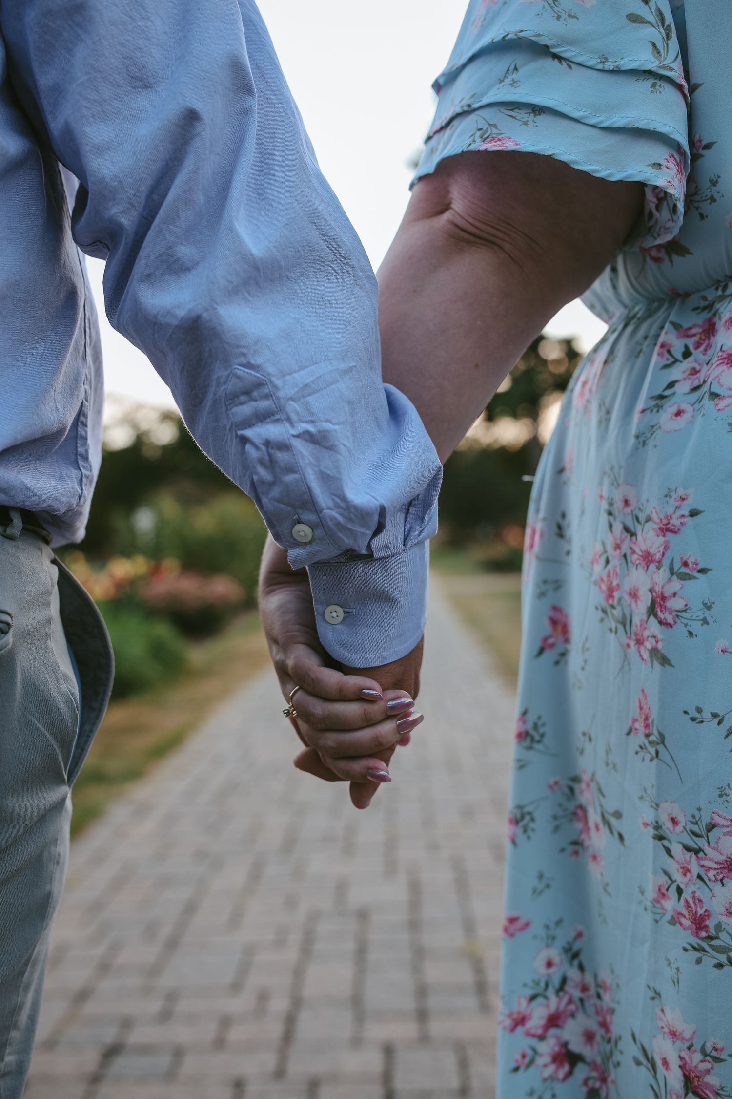 Close-up view of two people holding hands, one wearing denim and one in a floral dress, walking on a paved path.