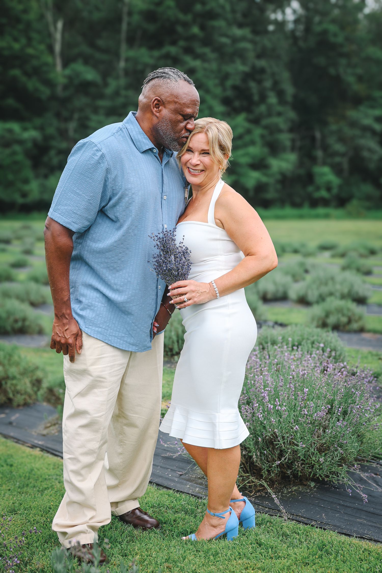 A couple embraces in a lavender garden, wearing a blue shirt and white dress on a summer day.