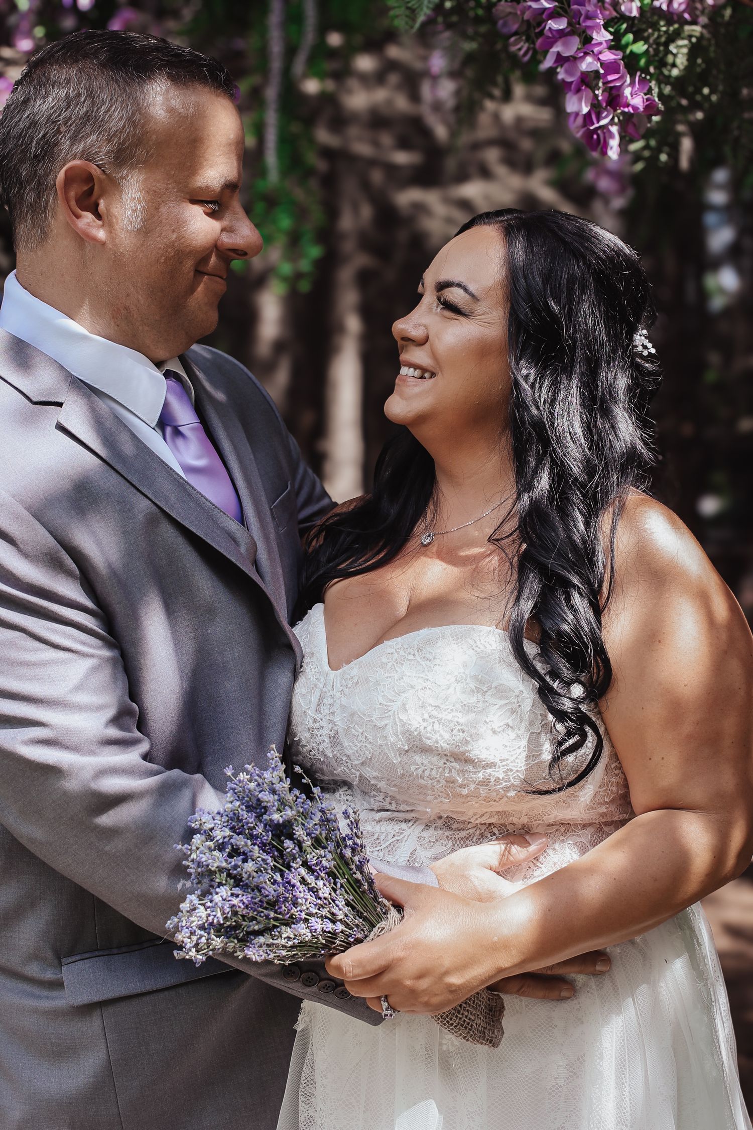 A romantic wedding couple in grey and white attire share tender moments against a backdrop of purple flowers.