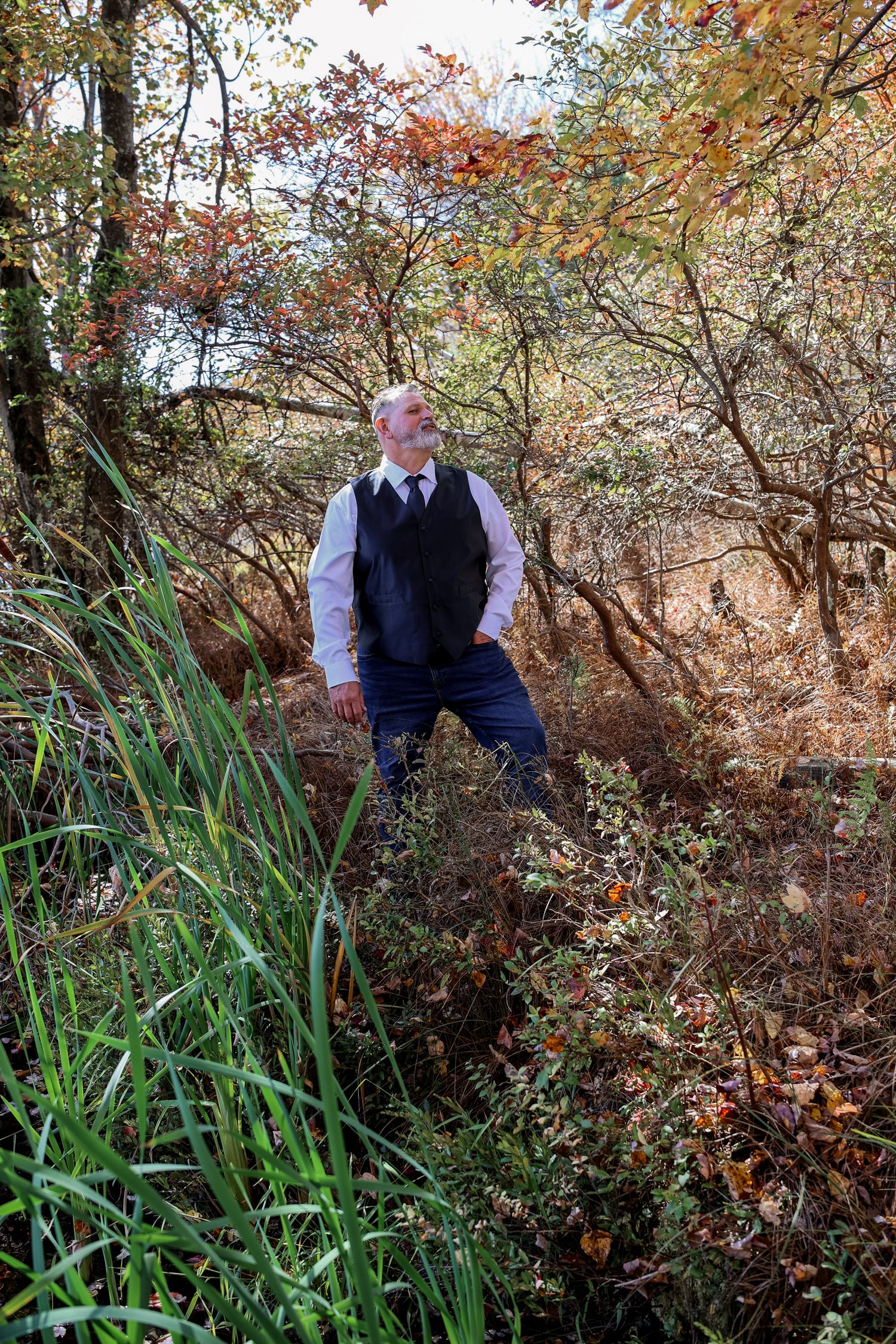 Person in blue vest and white shirt explores autumn woodland area with tall grass and fall foliage.
