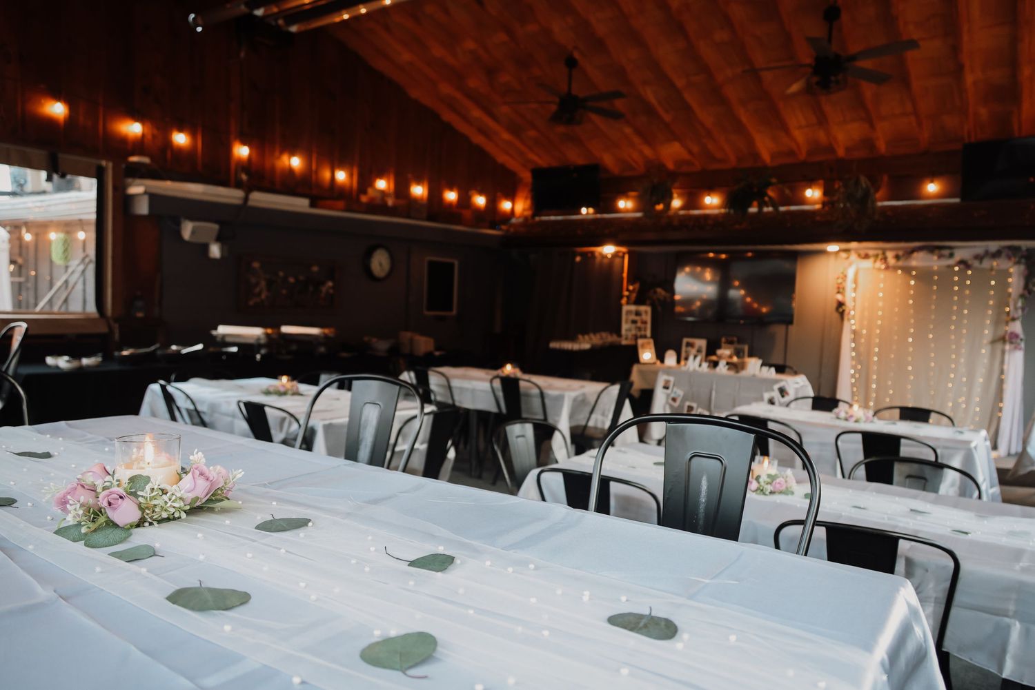 Wedding reception venue with rustic wooden ceiling, string lights, black chairs and white tablecloths decorated with floral centerpieces.