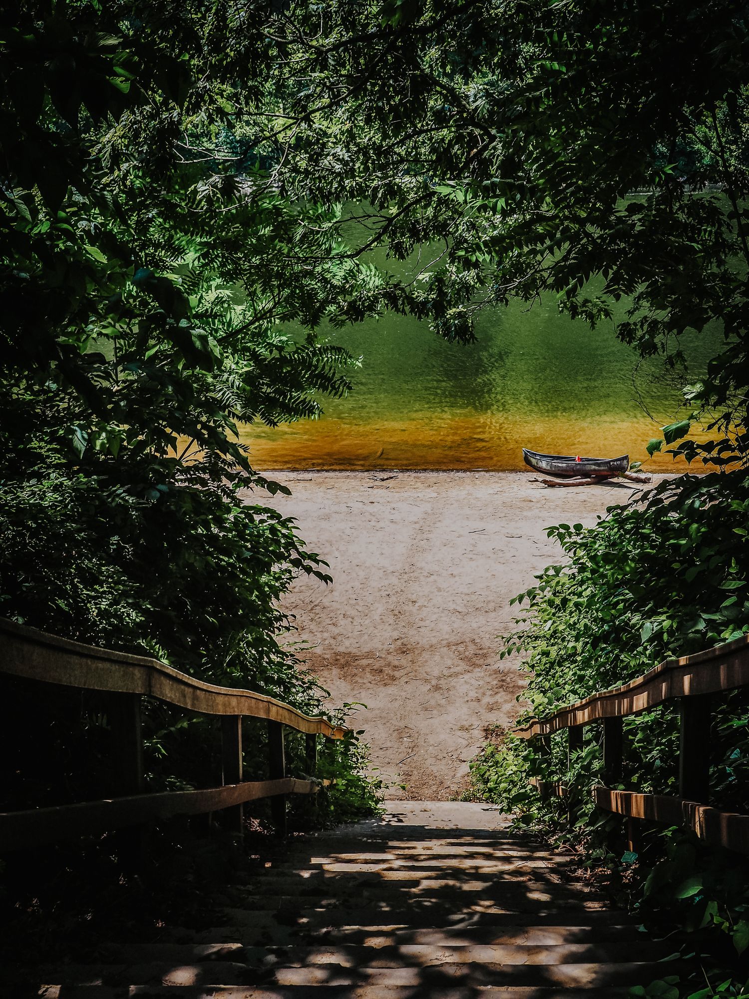Wooden boardwalk leads through lush green trees to a sandy beach and calm lake with golden ripples at sunset.