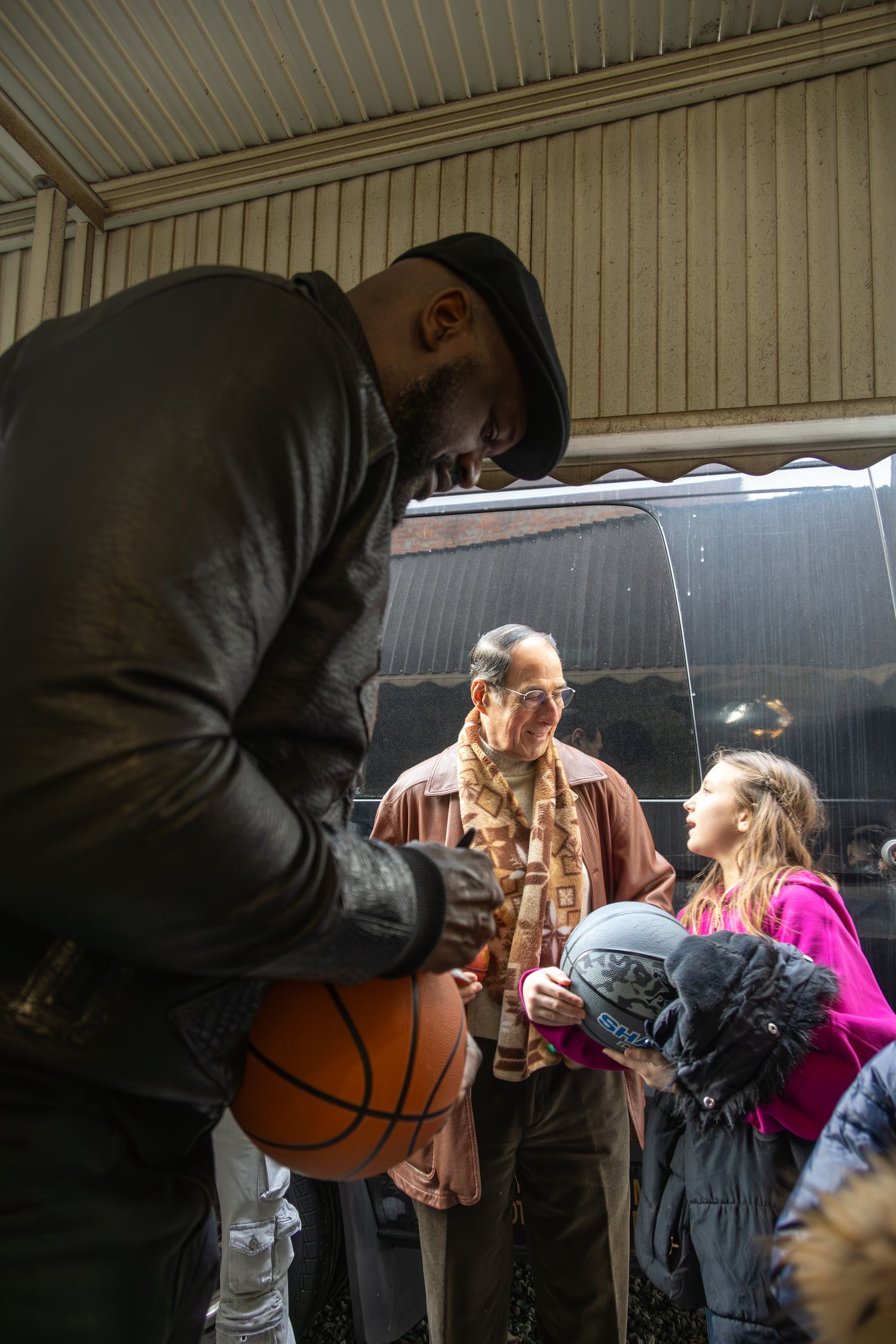 A person in a leather jacket holds a basketball while others gather around outside.
