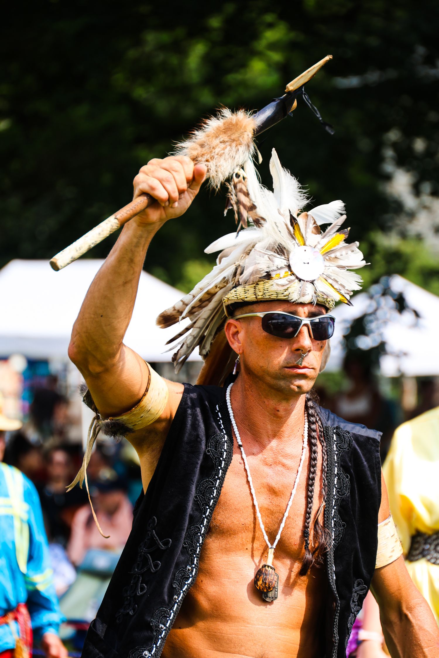 Shirtless festival-goer wearing feathered headdress and black vest raises ceremonial staff at outdoor gathering.