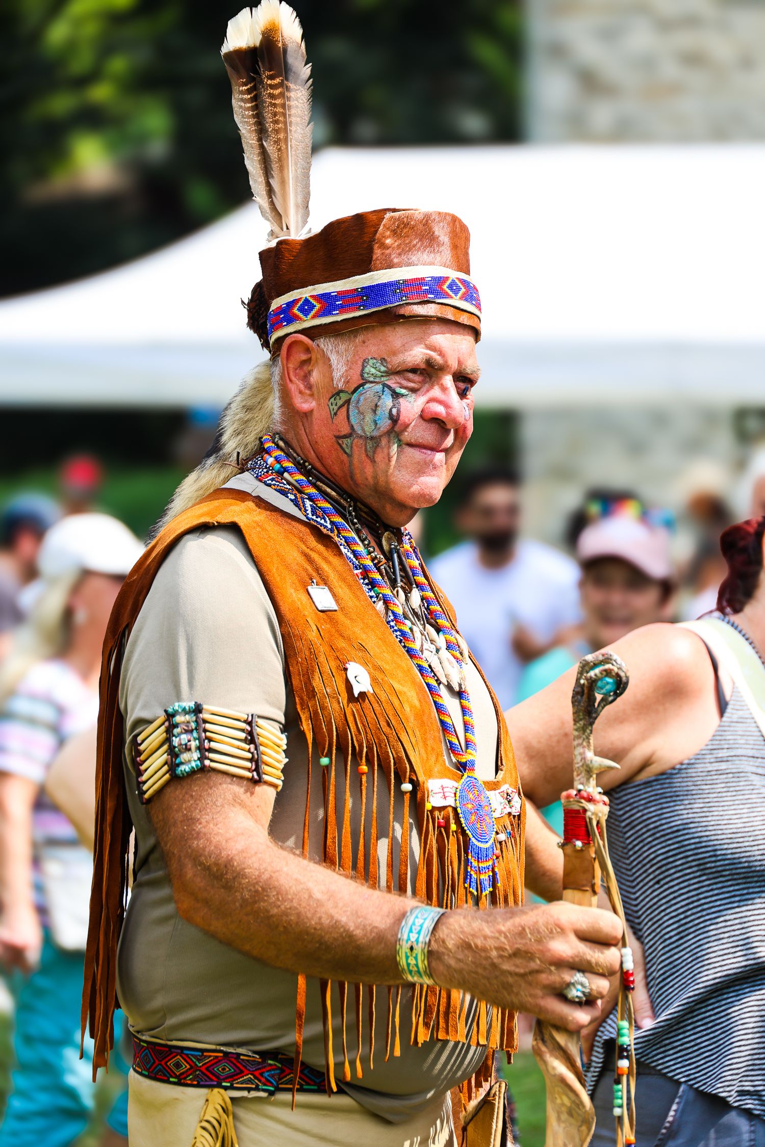 Native American performer in traditional fringed buckskin and face paint plays flute at outdoor cultural festival.