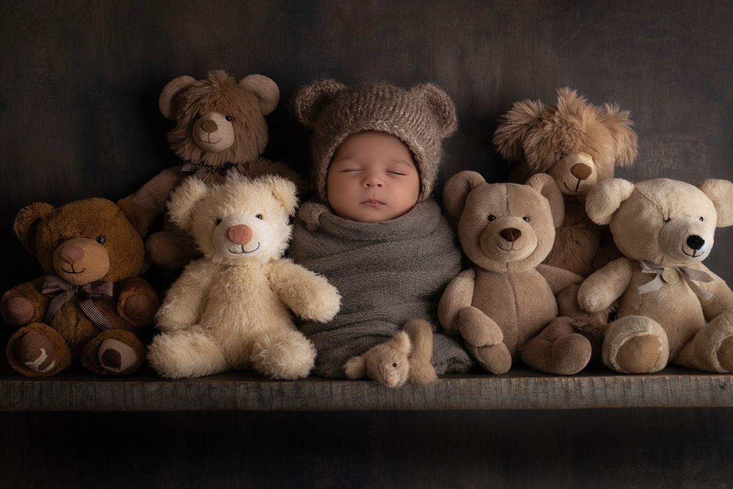 Baby in gray wrap and bear hat nestled among collection of teddy bears on dark background.