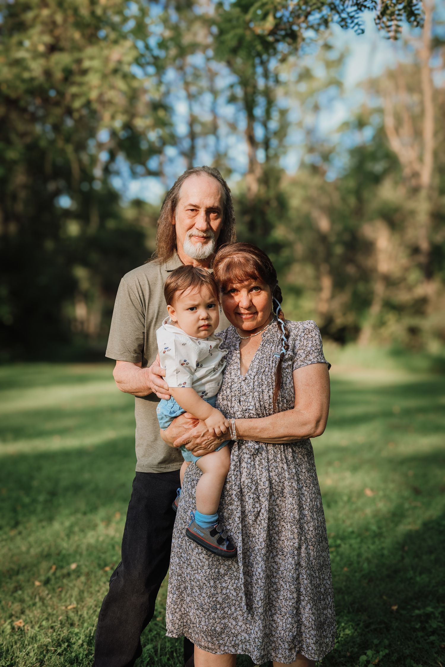 A family embraces together outdoors in a park with green trees and grass in the background during golden hour.