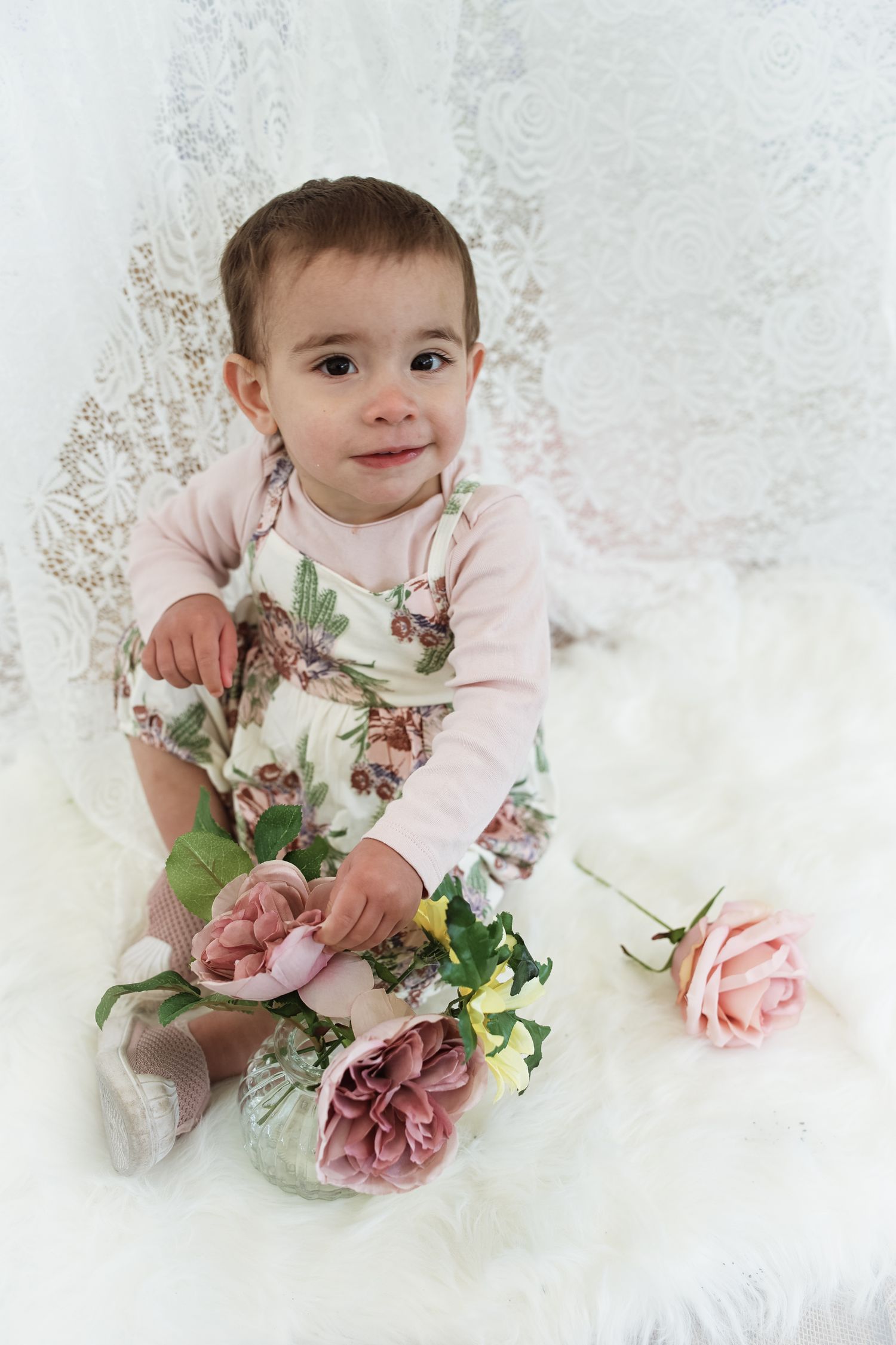 A child in pink sits on white fabric with delicate pink and purple flowers creating a dreamy portrait setting.