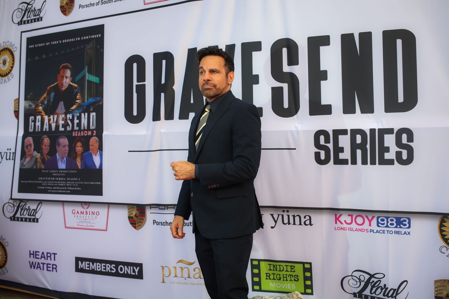 A person stands in front of a promotional backdrop with sponsor logos and text displays at a media event.