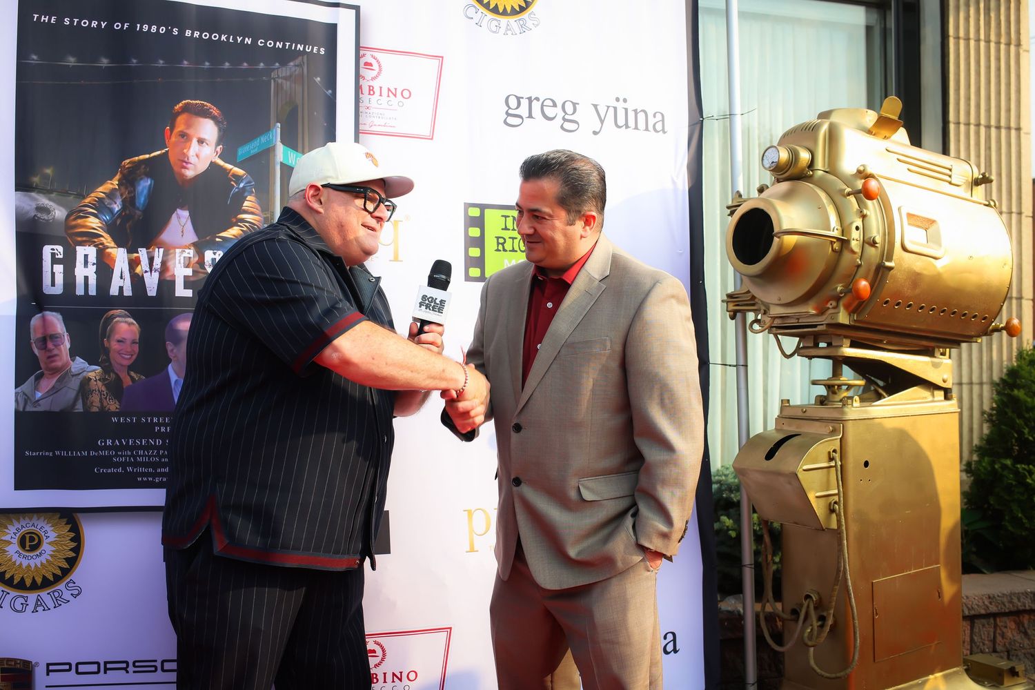 Two people shake hands at a movie premiere event with a film poster and golden camera prop in the background.