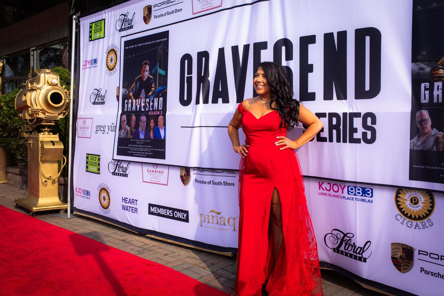 A red carpet event setup with a Gravesend series backdrop banner and someone posing in a long red gown.