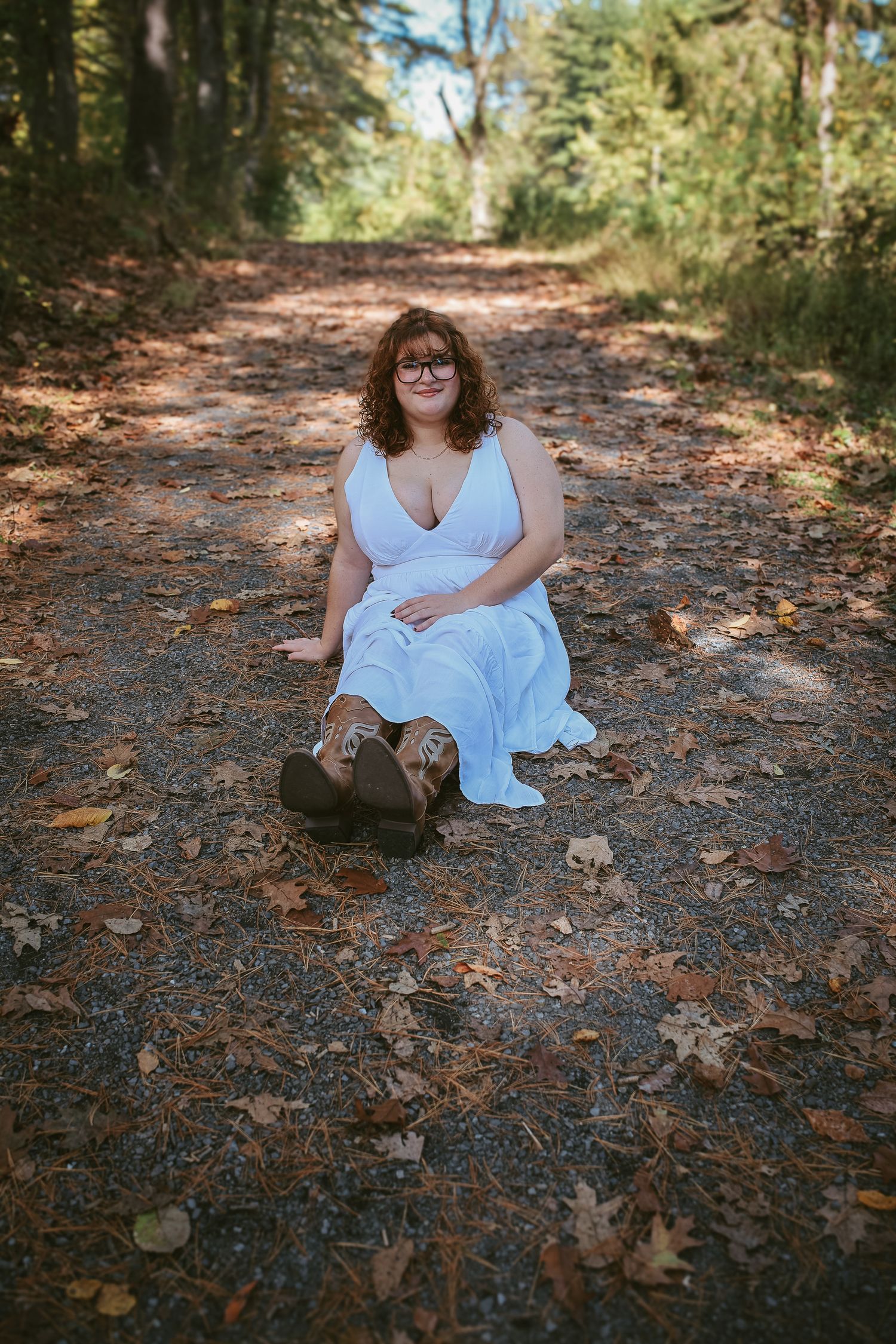 Person in a light blue dress sits on a forest path covered with fallen autumn leaves.