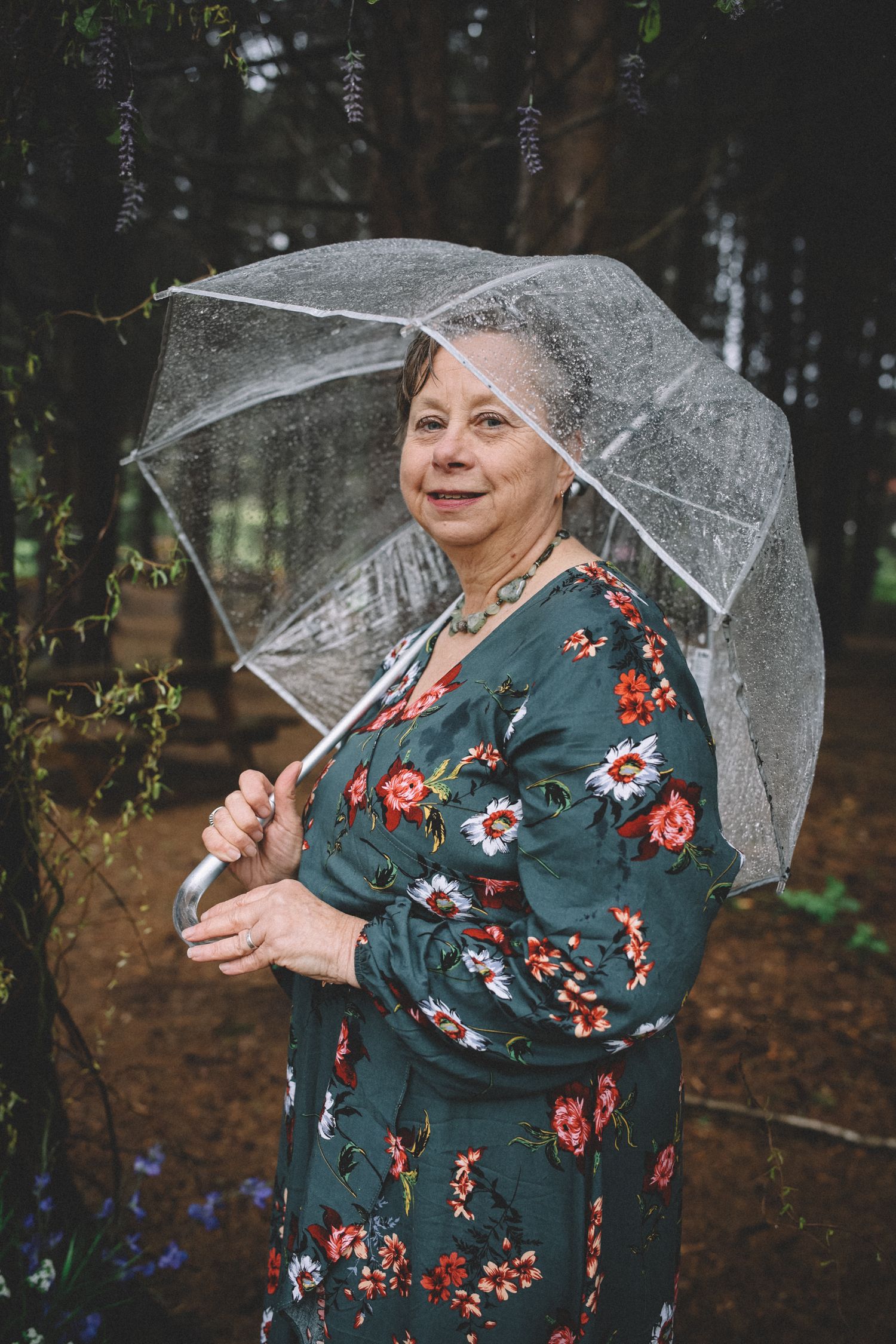 A person in a floral dress holds a clear umbrella in a moody forest setting.