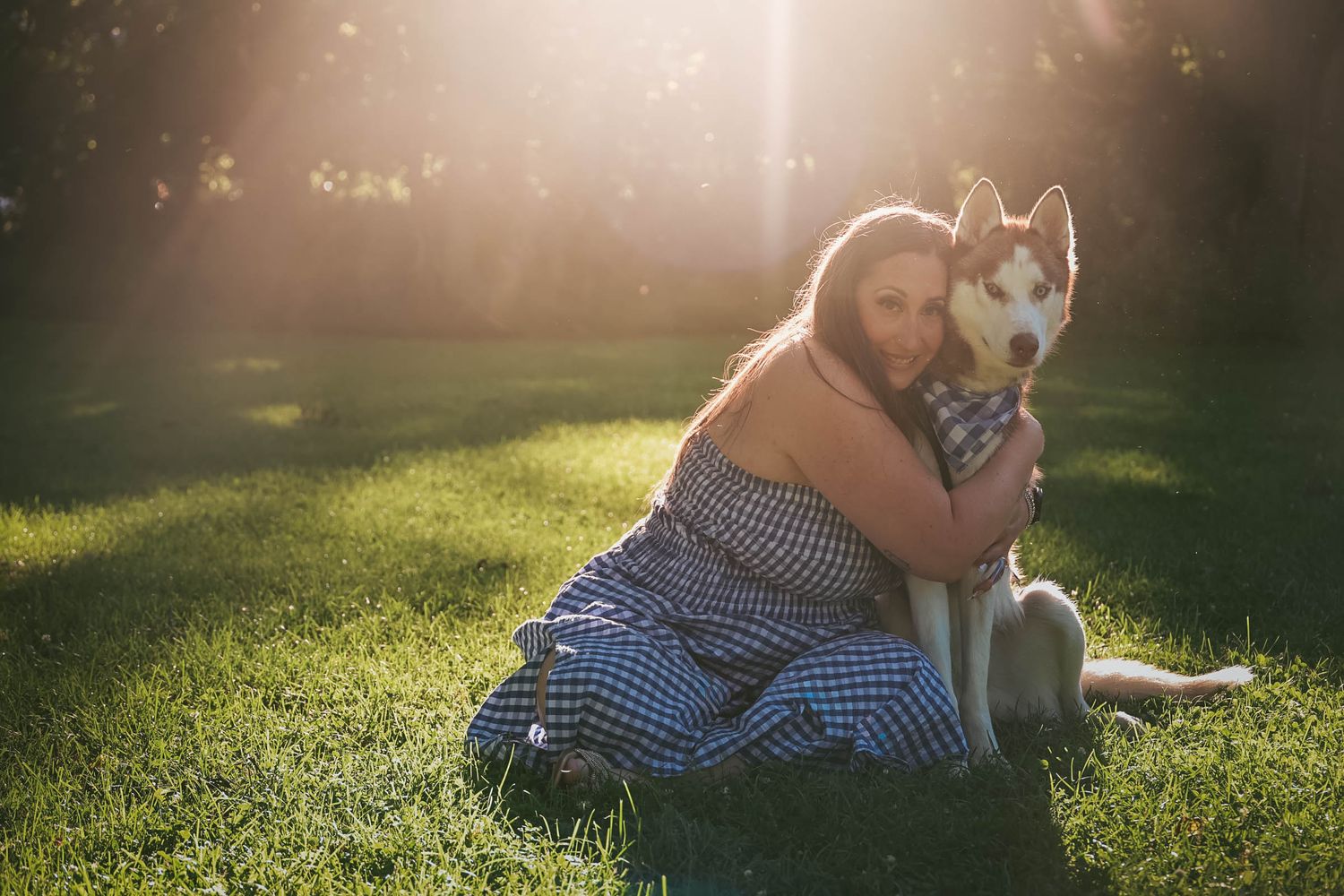 Woman hugging Siberian Husky dog on green grass in warm sunlit park during golden hour.