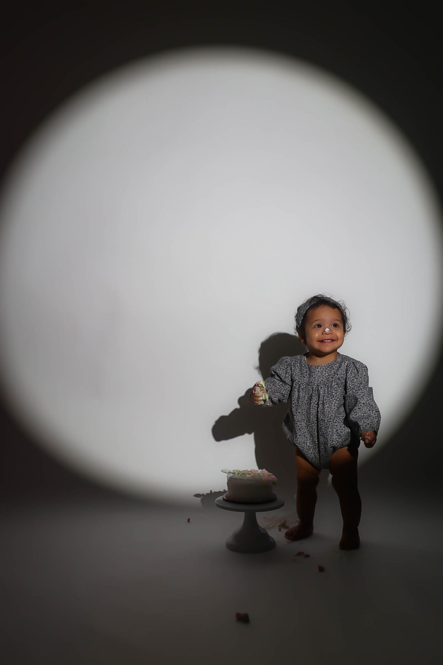 Smiling toddler in gray romper standing beside a small cake under a circular spotlight on a dark background.
