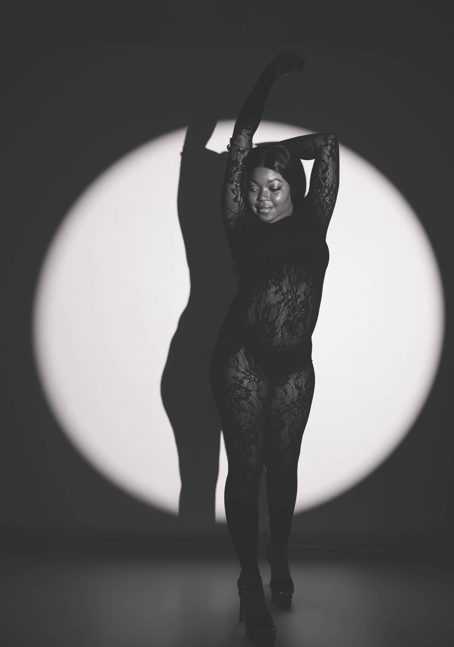 Black and white photo of a woman in lace bodysuit posing gracefully with arms raised against a circular spotlight backdrop.