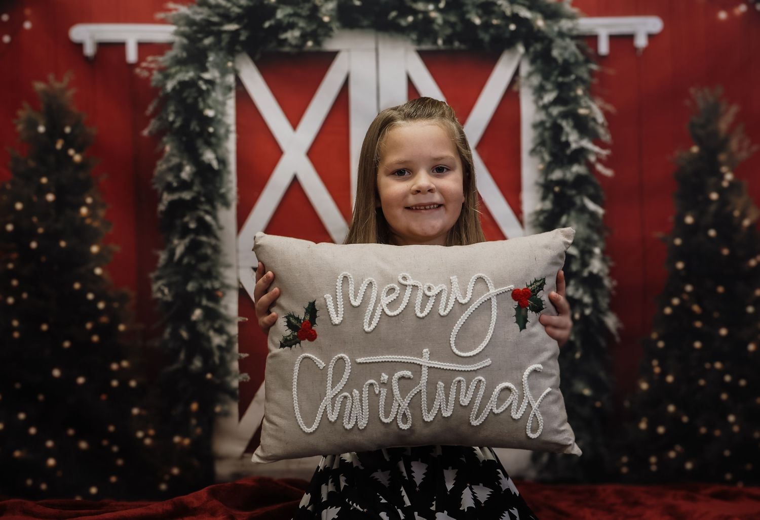 Young girl smiling and holding a Merry Christmas decorative pillow in front of a festive red barn holiday backdrop.
