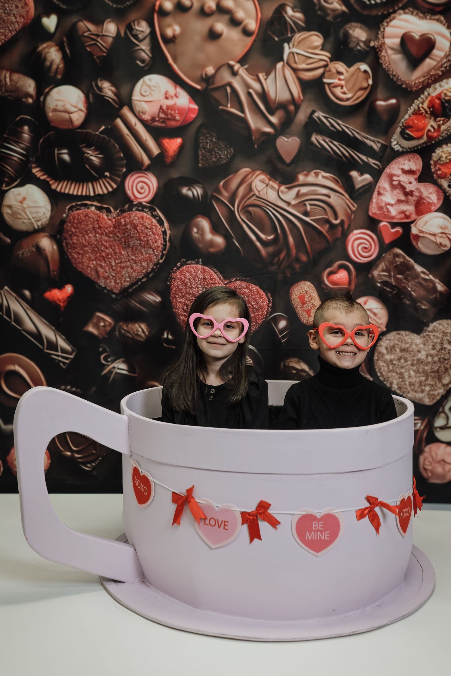 Two kids in heart glasses sit in a giant teacup with Valentine candy backdrop for a festive photo.