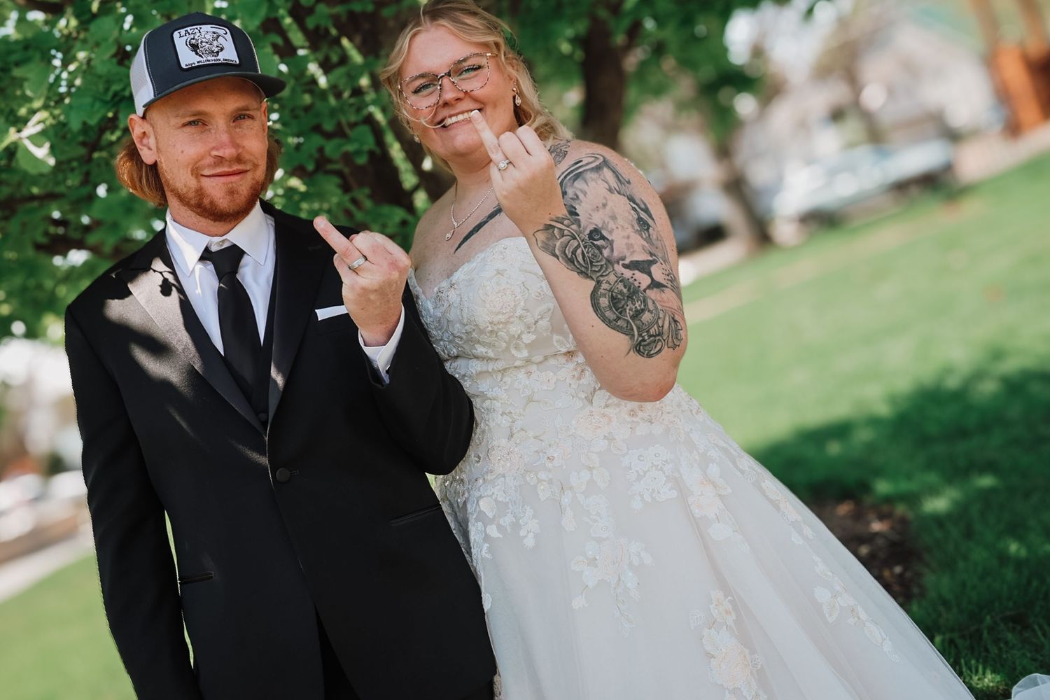 Newlywed couple in wedding attire posing playfully outdoors, bride in lace gown with tattoos, groom in black suit.