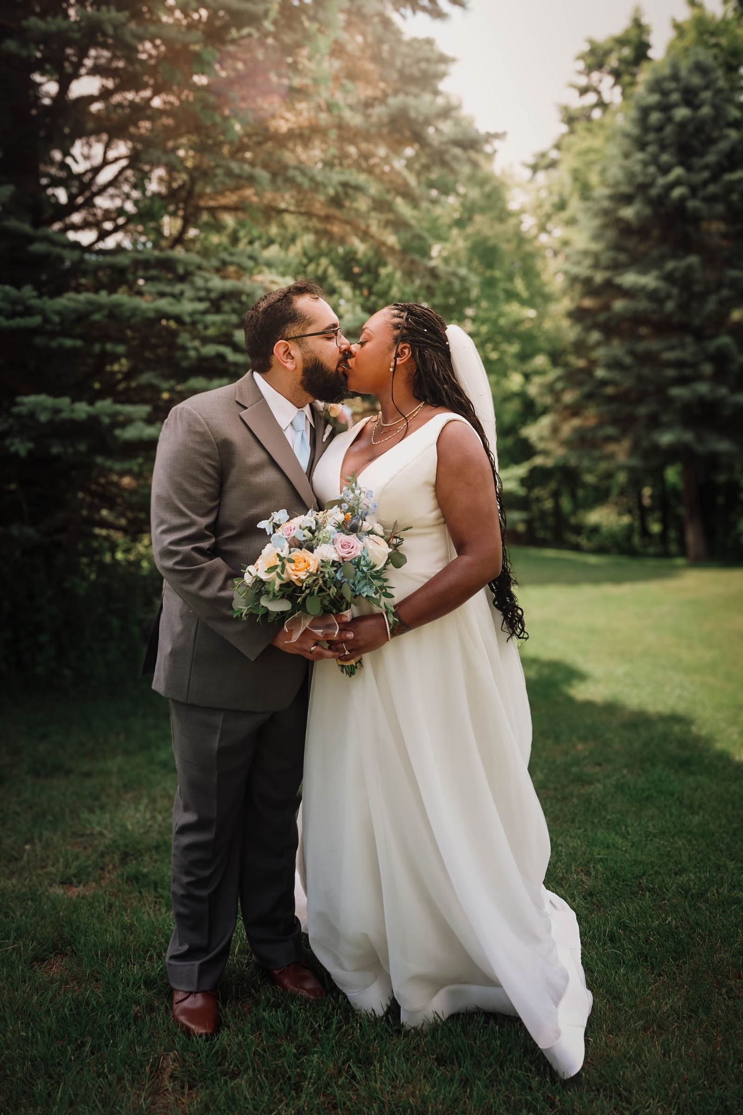 Bride and groom sharing a kiss outdoors, bride in white gown holding floral bouquet, groom in gray suit, lush green trees behind.