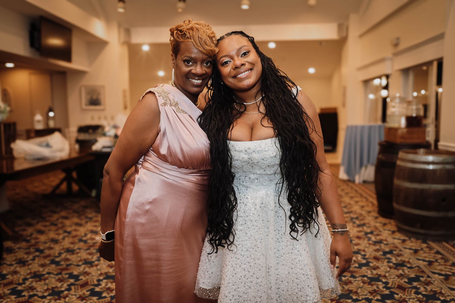 Two women in formal attire smile together at an elegant wedding reception venue.