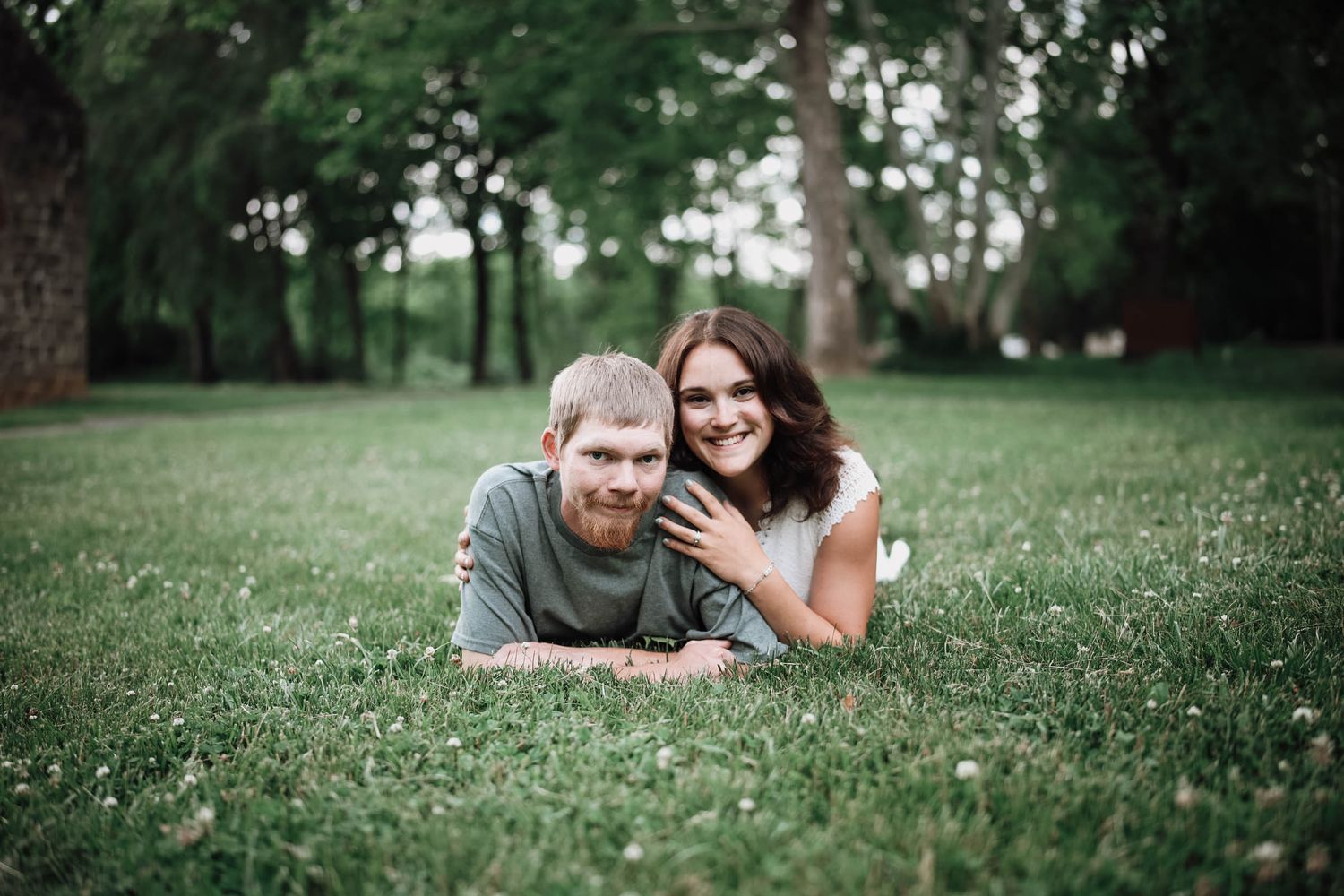 Couple laying on green grass in a park, smiling at camera with trees in background.
