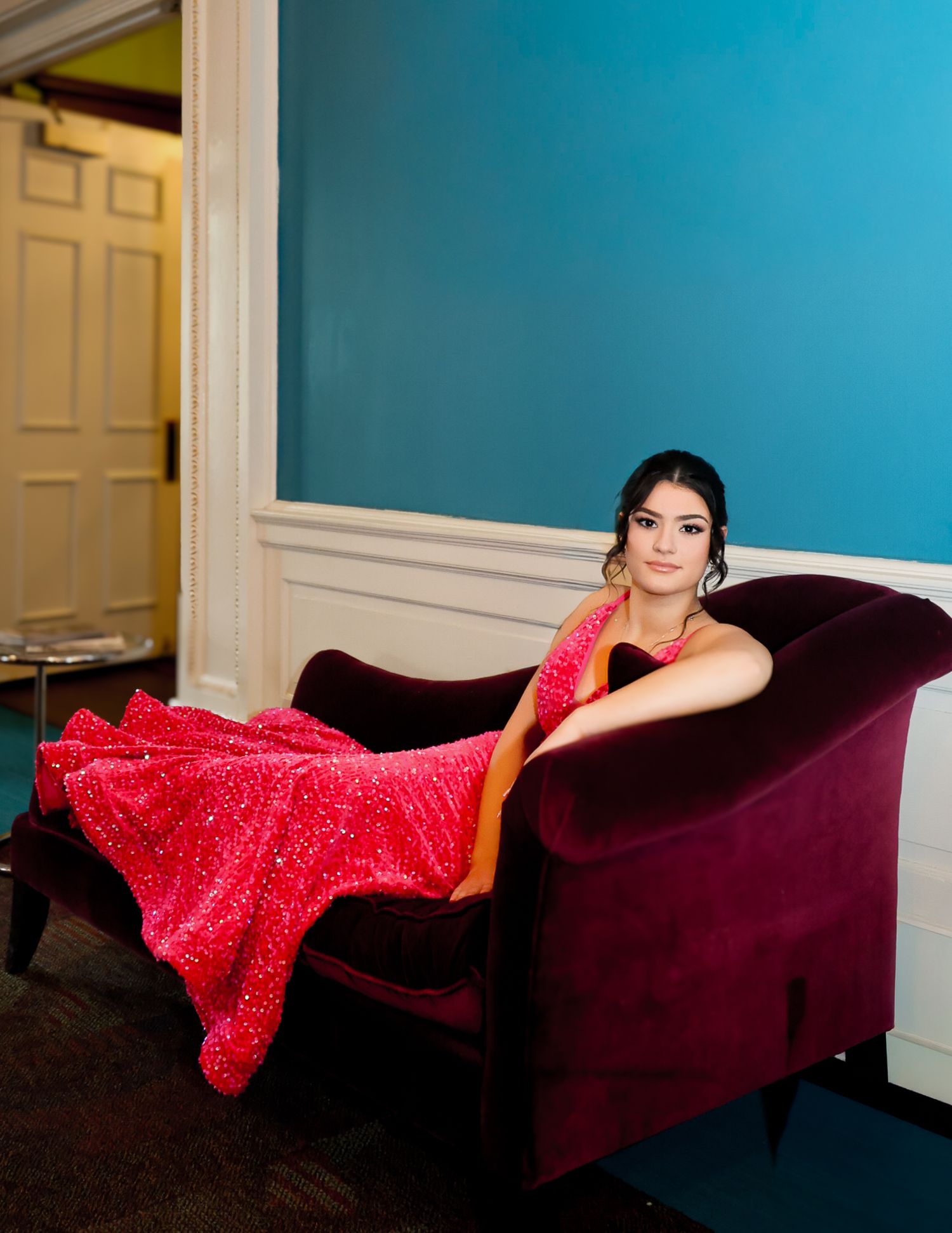 Young woman in a sparkling red gown poses elegantly on a velvet chaise lounge against a teal wall.