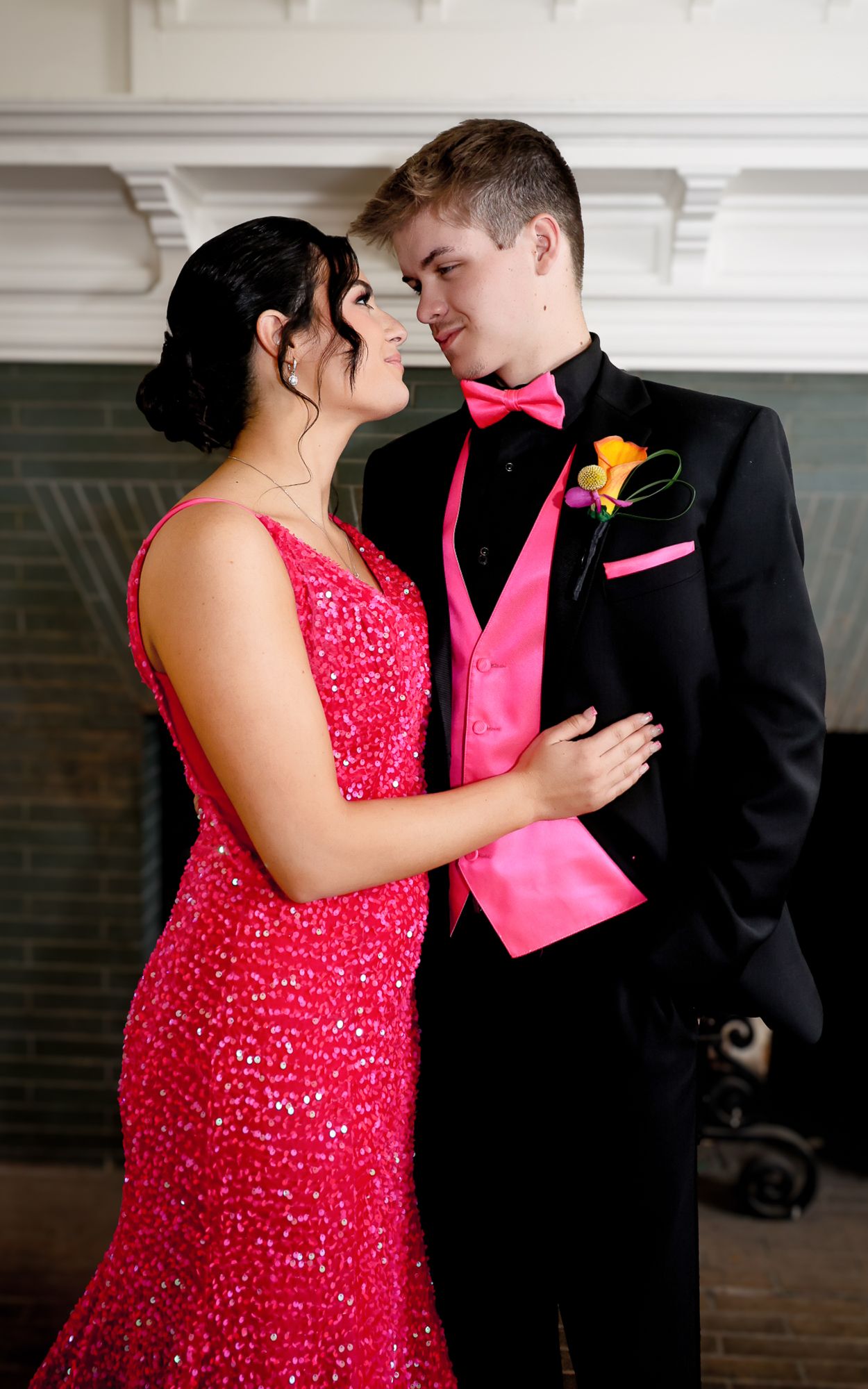 A couple dressed in matching hot pink prom attire, with the girl in a sequined gown and the boy in a black tuxedo, share a romantic moment.