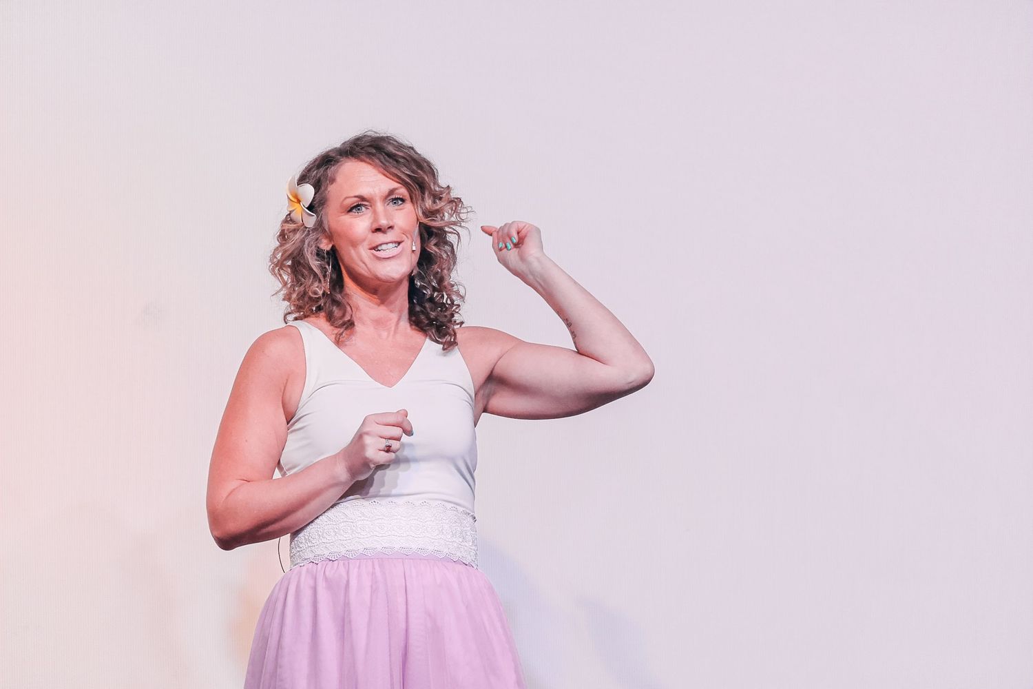 Woman in white tank top and pink tulle skirt flexing her arm muscle while smiling confidently.