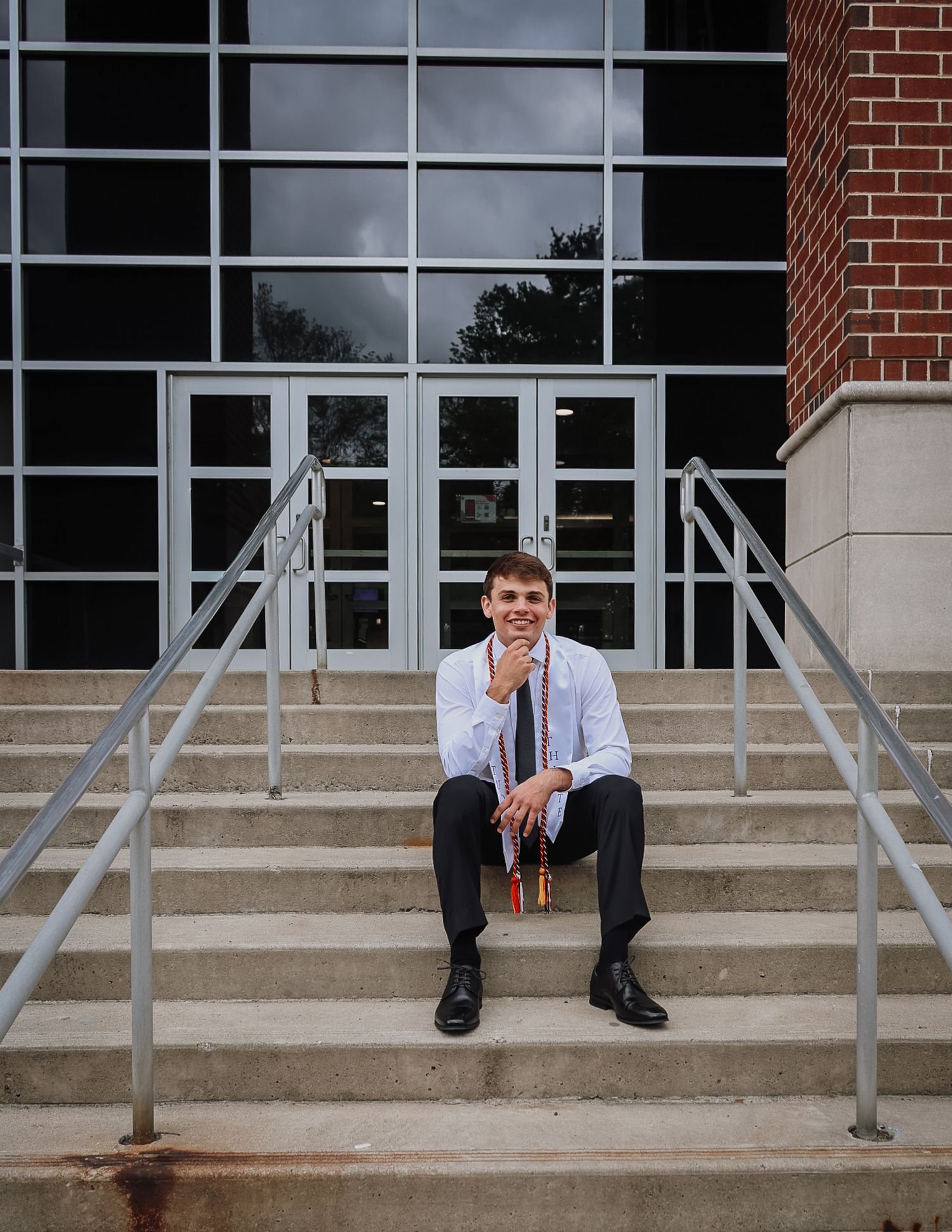 Young man in dress shirt and tie sits on university building steps holding graduation regalia, smiling confidently.
