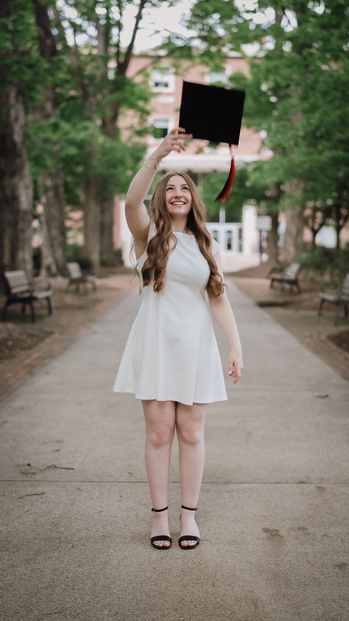 Smiling young woman in white dress holding graduation cap overhead on a tree-lined campus walkway.