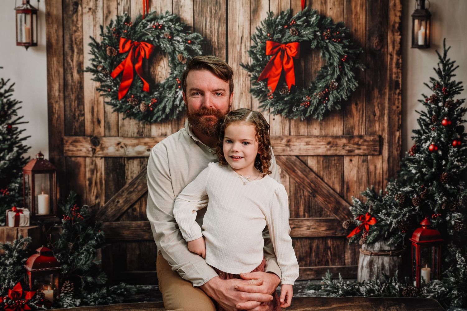 Two people pose in front of rustic wooden doors decorated with Christmas wreaths, trees and holiday decor.