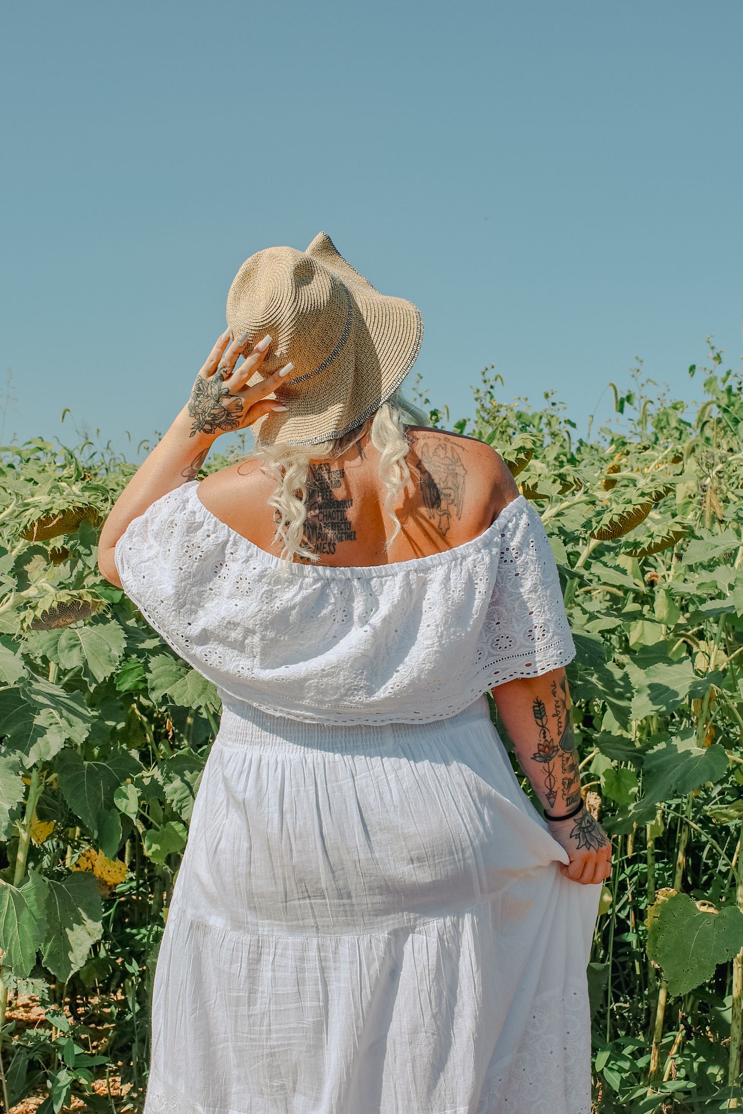A person in a white summer dress and straw hat stands in a lush green field of growing crops on a sunny day.