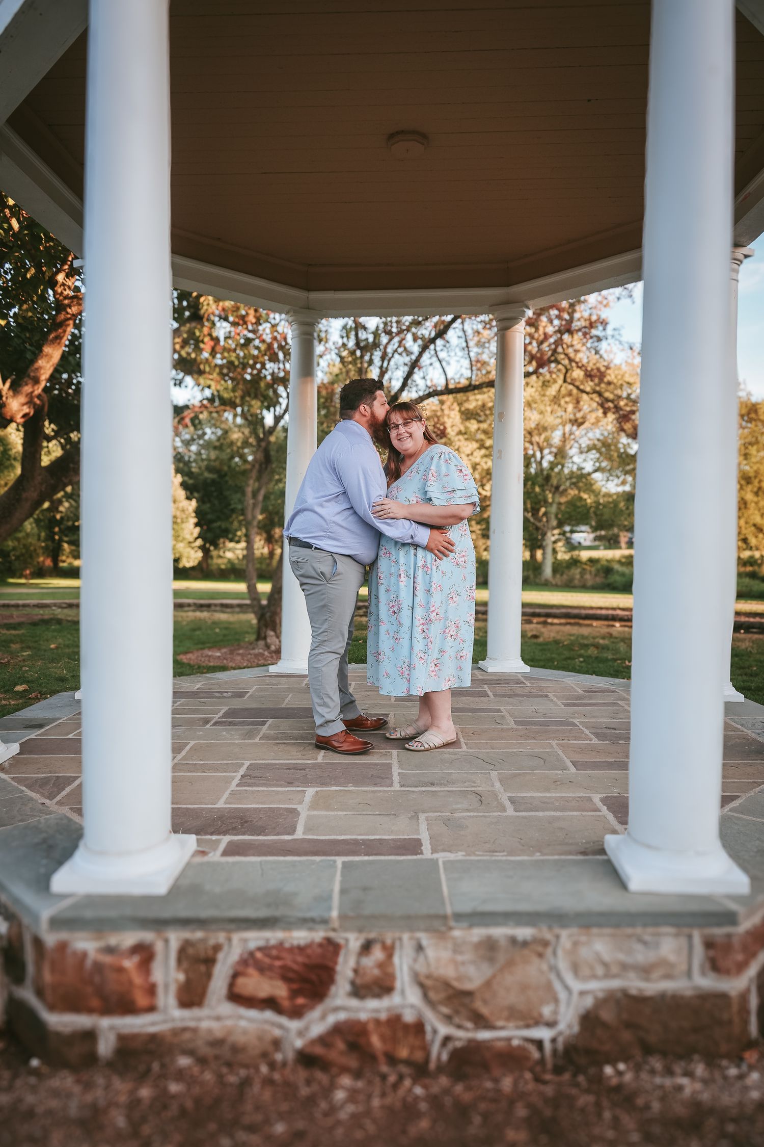 Two people share a tender moment under a white columned gazebo on a stone patio during golden hour.