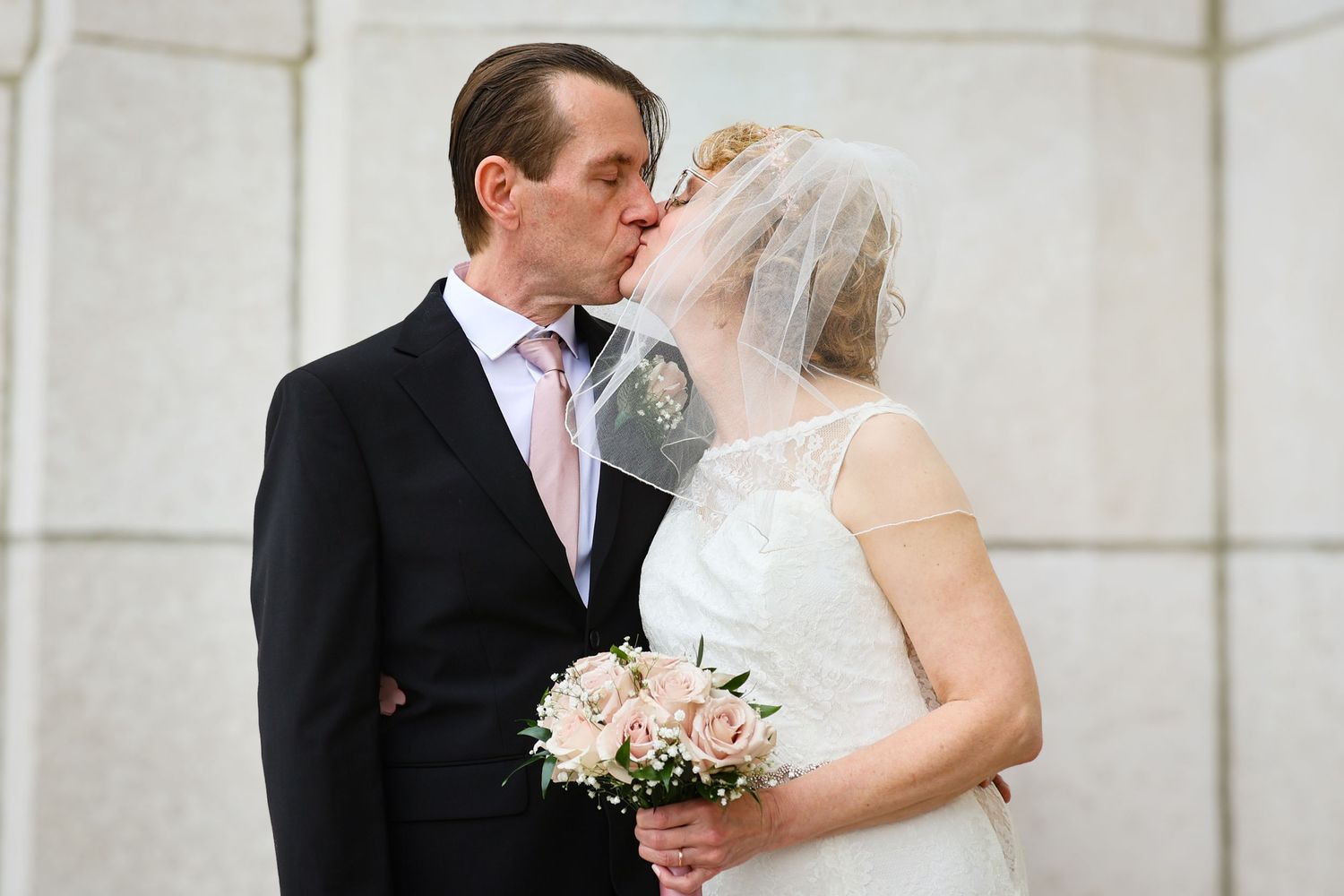 A romantic wedding moment captured outside a white stone building with a bridal bouquet of white flowers.
