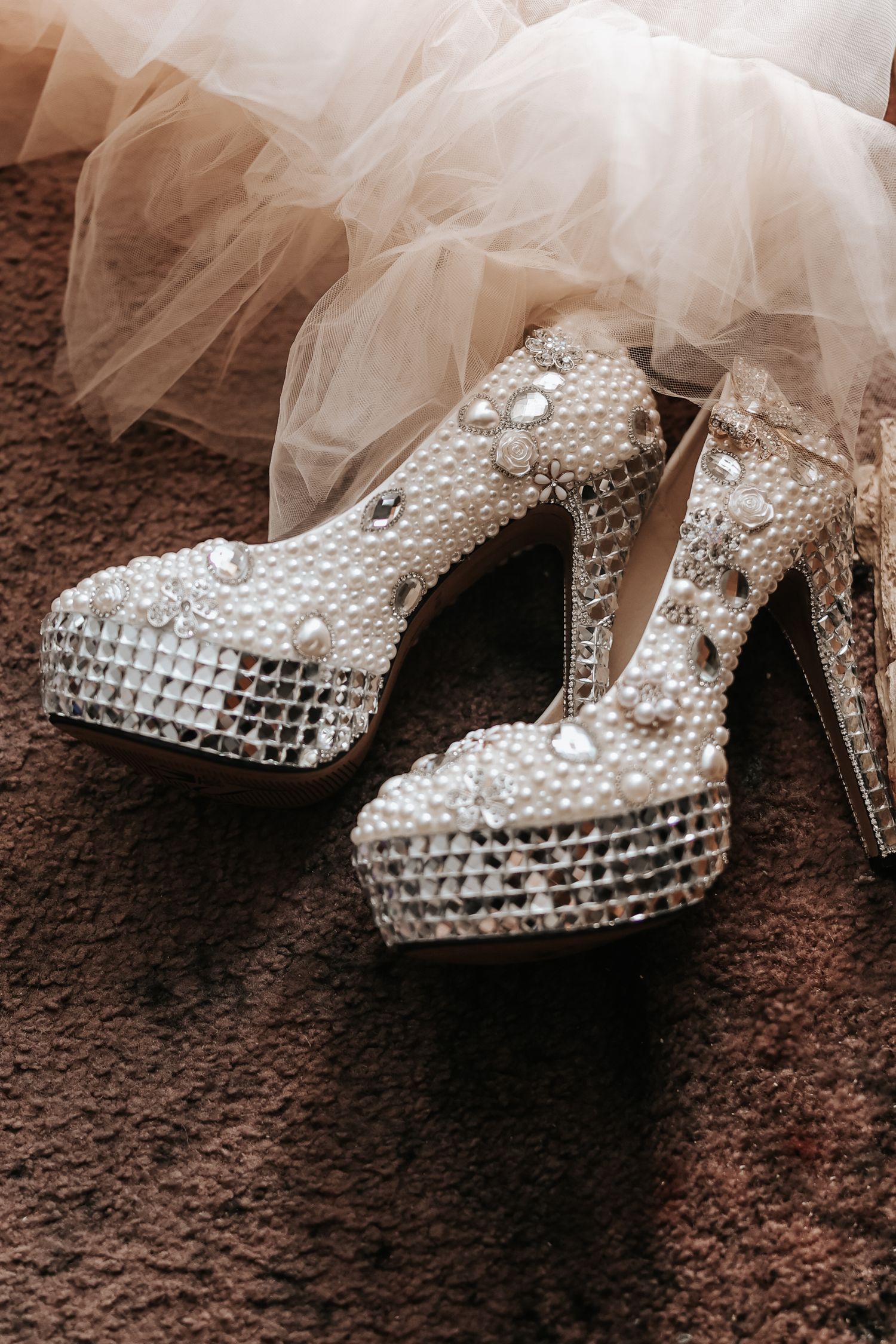 Sparkling crystal-encrusted high heel wedding shoes with platform soles next to white tulle veil on brown carpet.