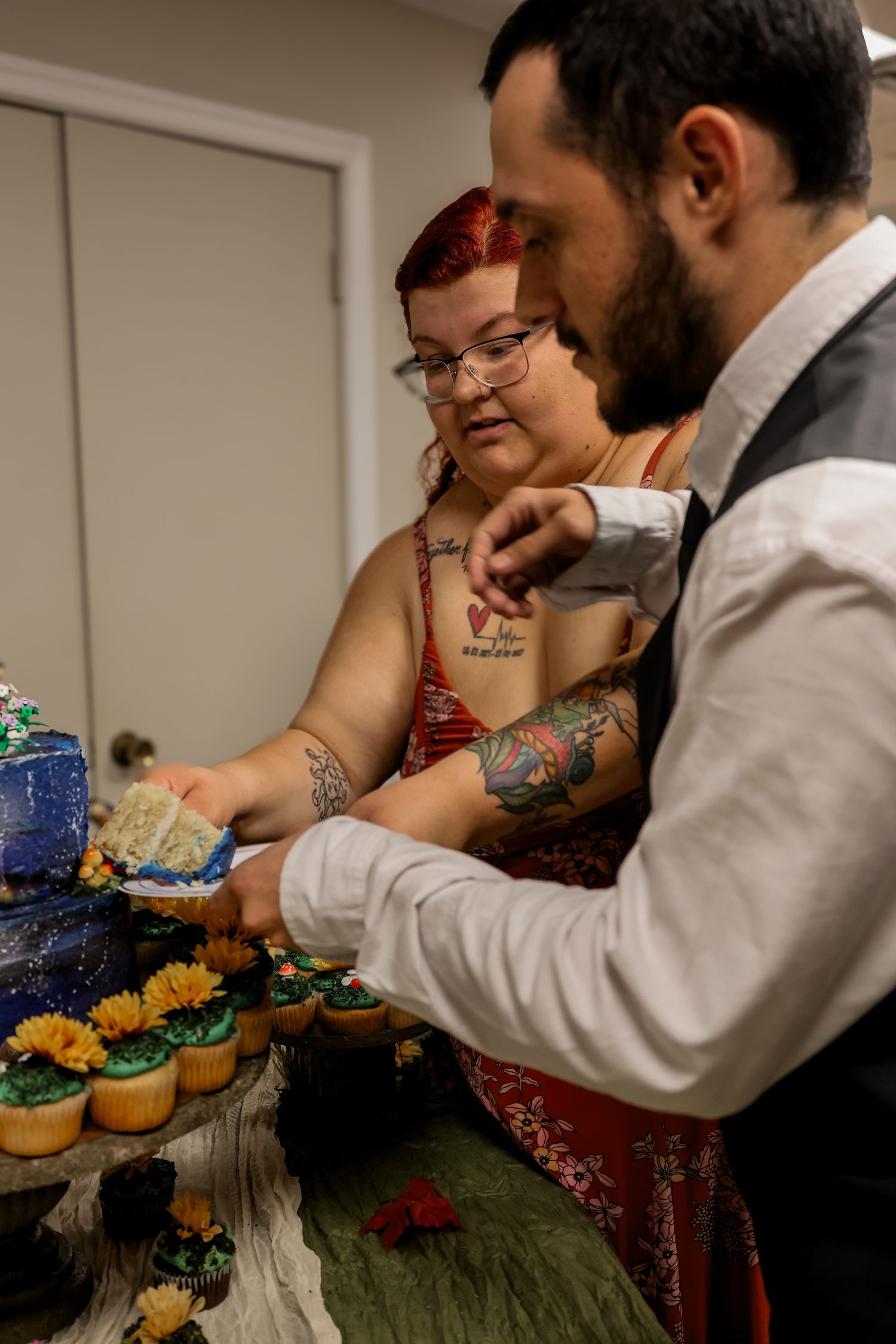 A couple cuts into their wedding cake together while standing at a dessert table decorated with colorful cupcakes.