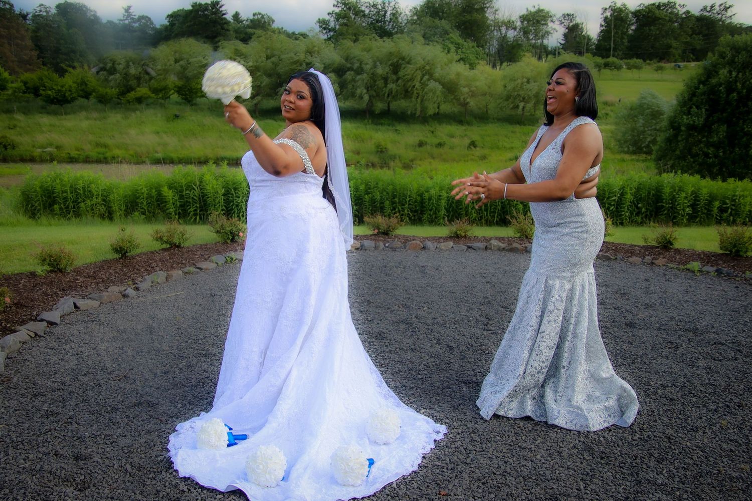 Two people in white wedding dresses pose playfully outdoors on a gravel path with green scenery in the background.