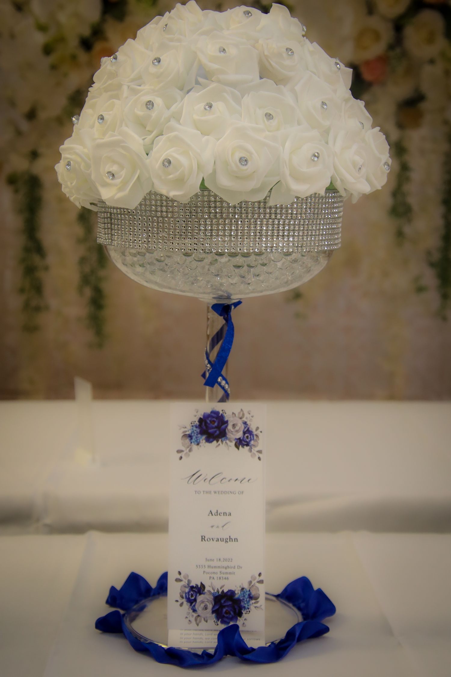 White rose centerpiece with royal blue ribbon accents and crystal details displayed at elegant wedding reception table setting.