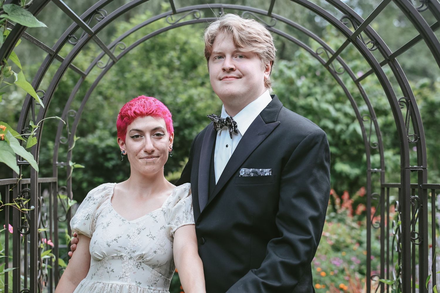 A couple poses together under a garden arbor, with the person on the left wearing pink hair and a lace dress.