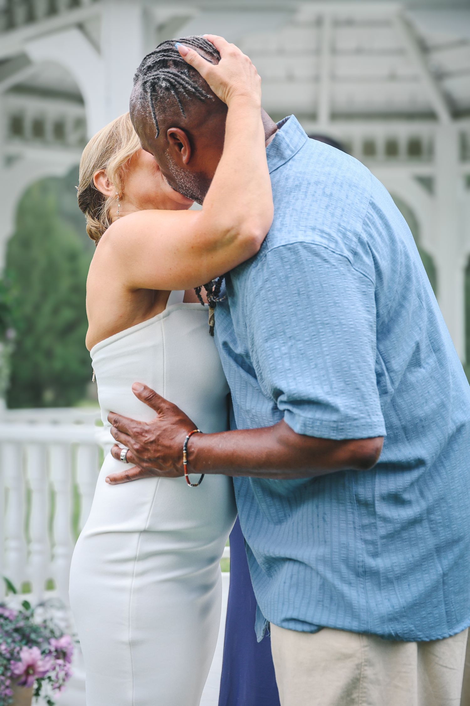 A couple in blue and white attire share a romantic kiss in front of a white picket fence and garden setting.