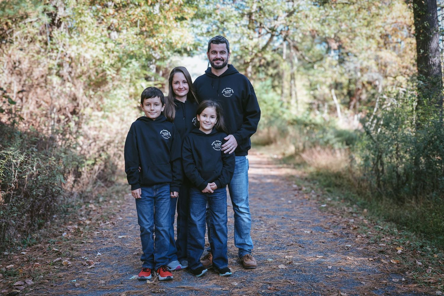 Family group poses together on a leaf-covered autumn trail through a wooded area.