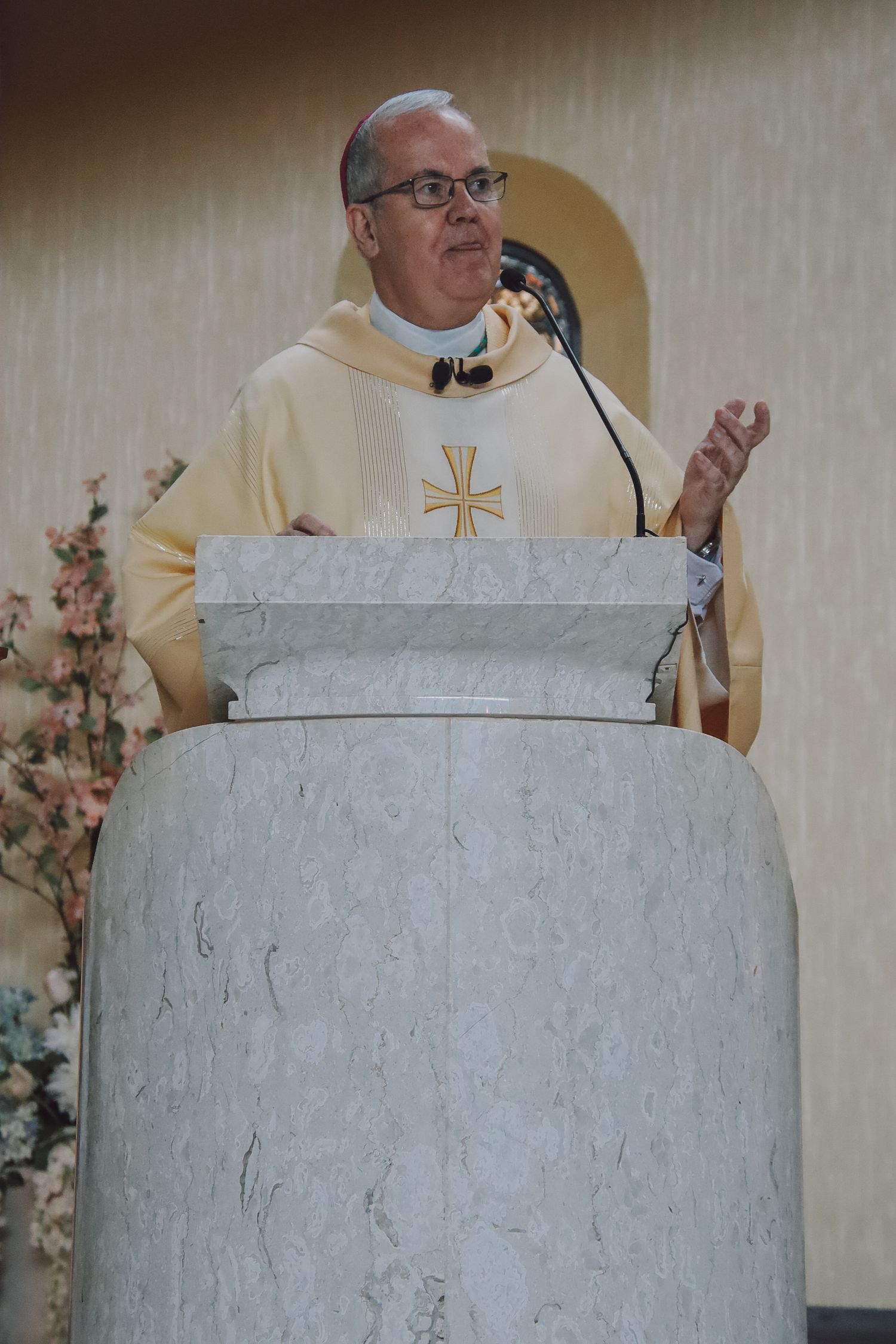 A priest in cream-colored vestments stands behind a white marble pulpit during a church service.