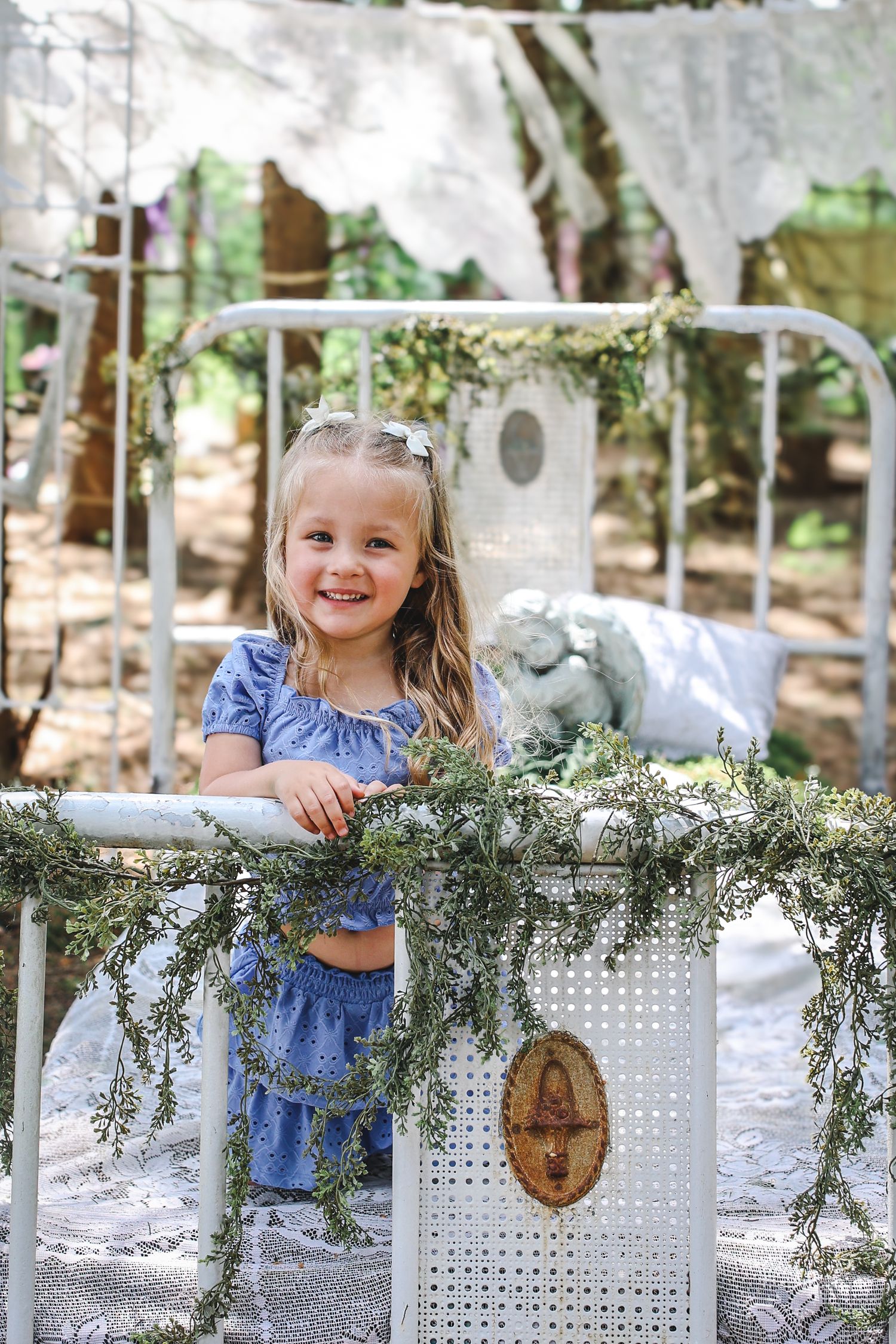 A child in a blue dress sits at a white table decorated with greenery garland for an outdoor party setting.