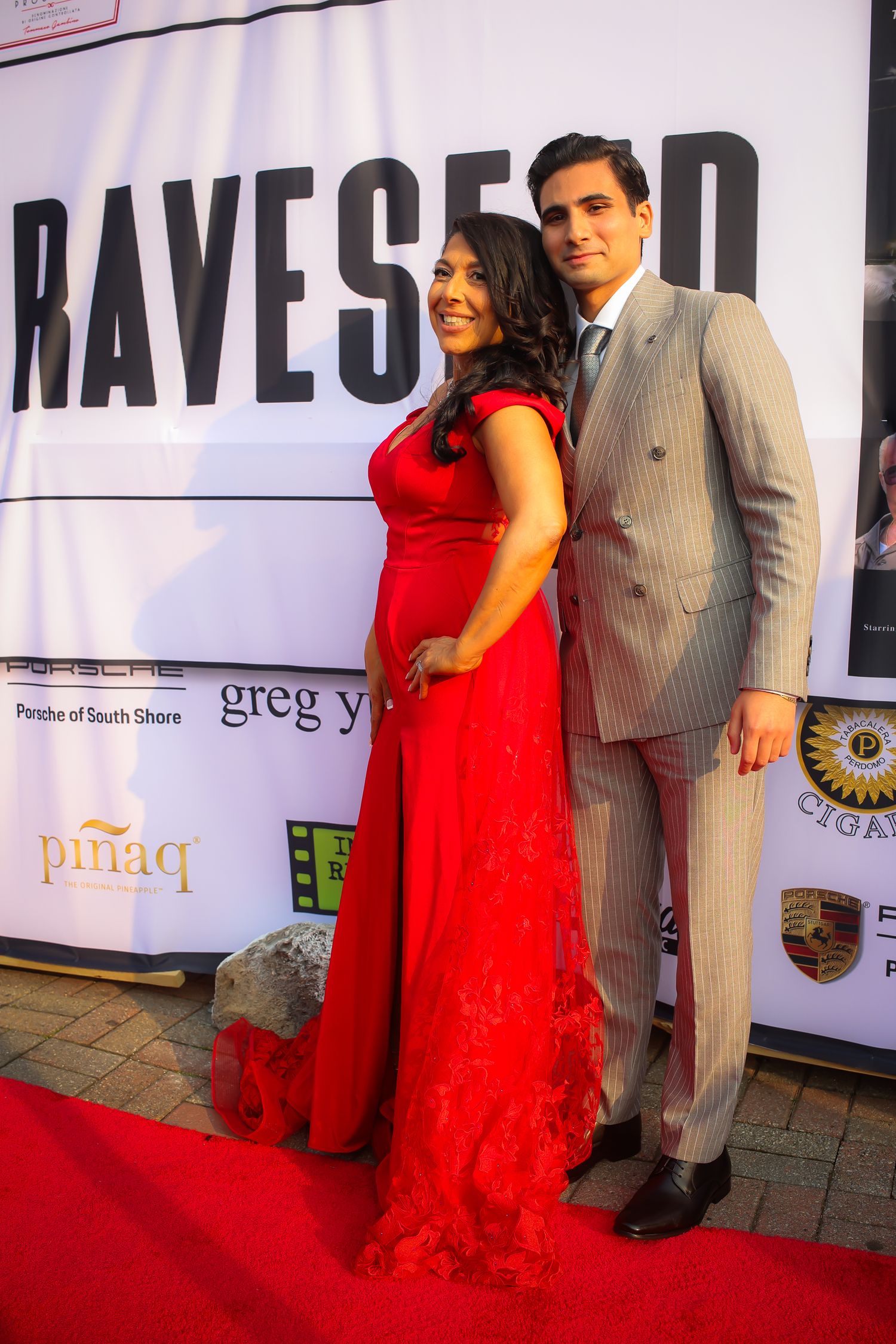 A couple poses on a red carpet event, with the woman wearing a flowing red dress and the man in a gray suit.
